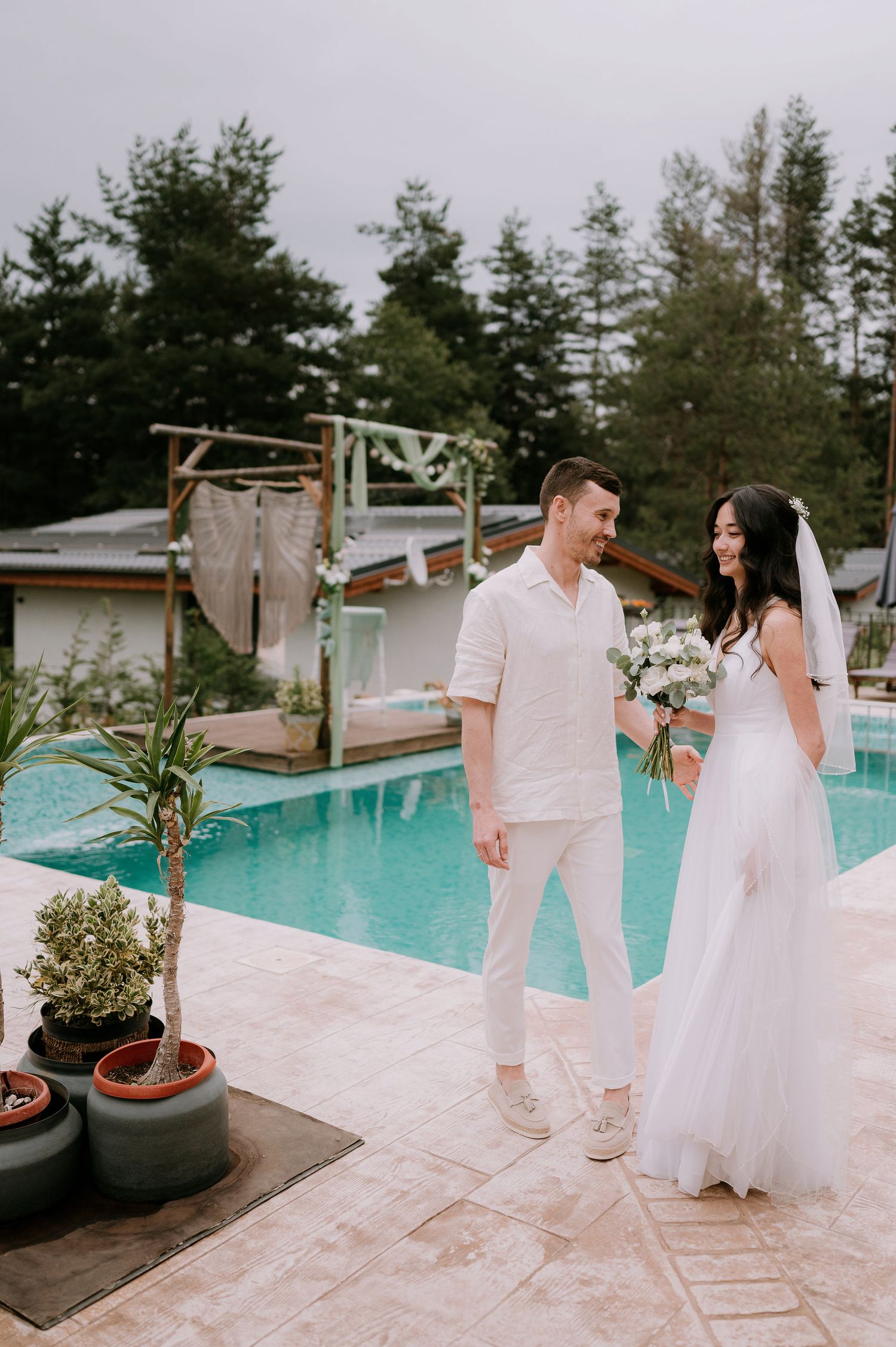 Wedding couple in white shares candid moments by a resort pool surrounded by pine trees.