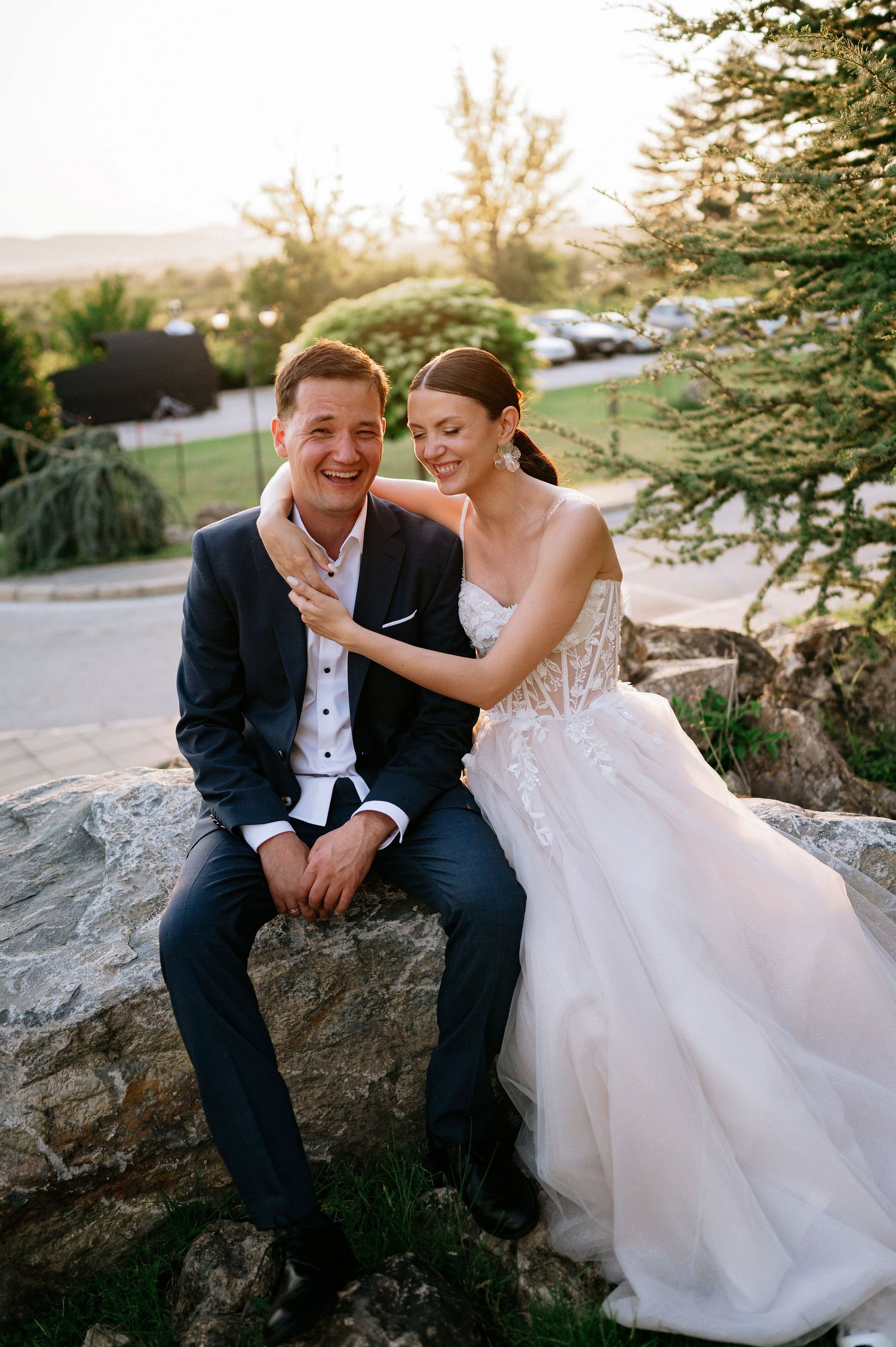 Happy wedding couple enjoys playful moment on stone garden feature at sunset.