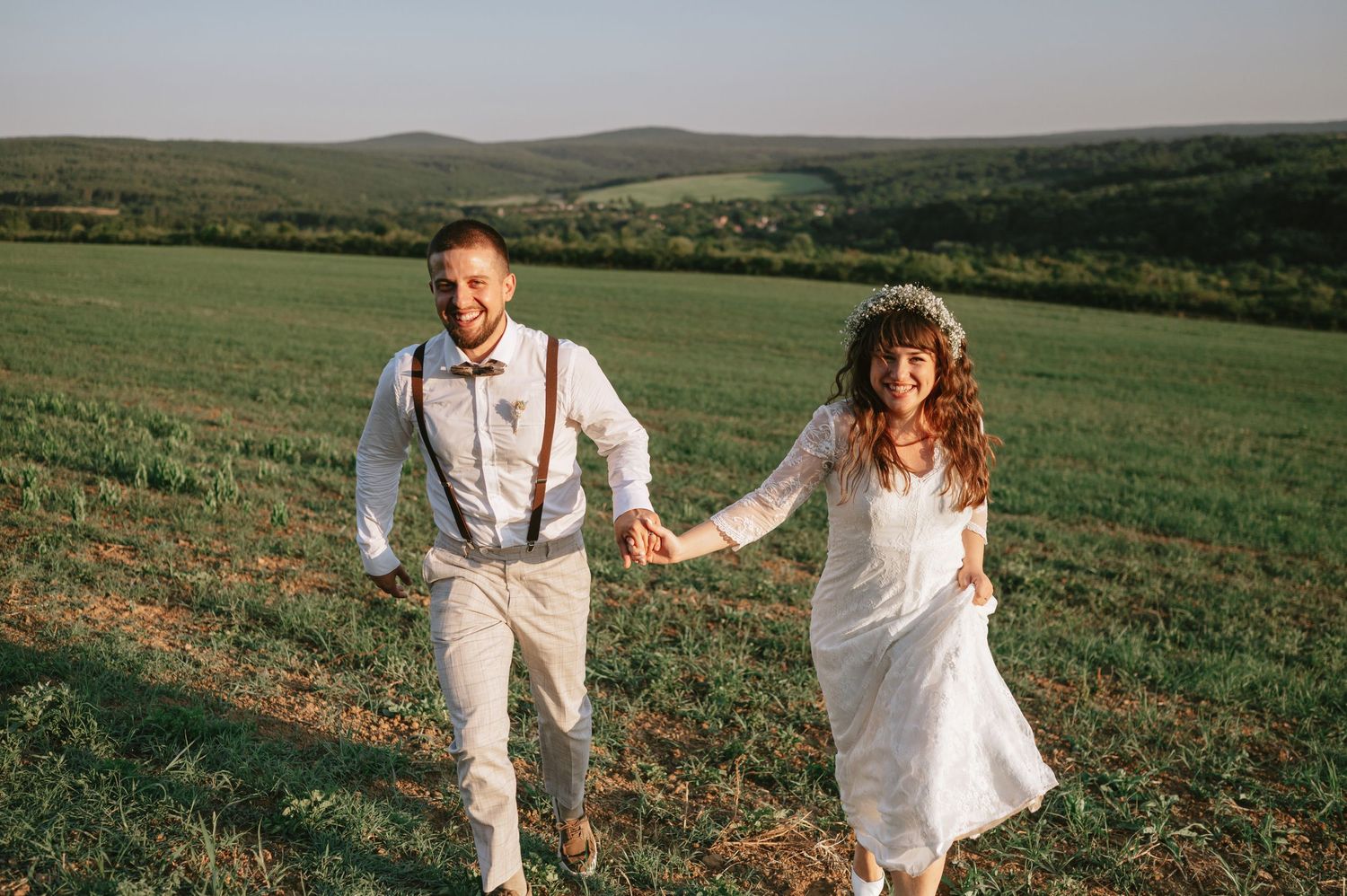 Two people in white outfits hold hands while walking across a scenic grassy hilltop landscape.