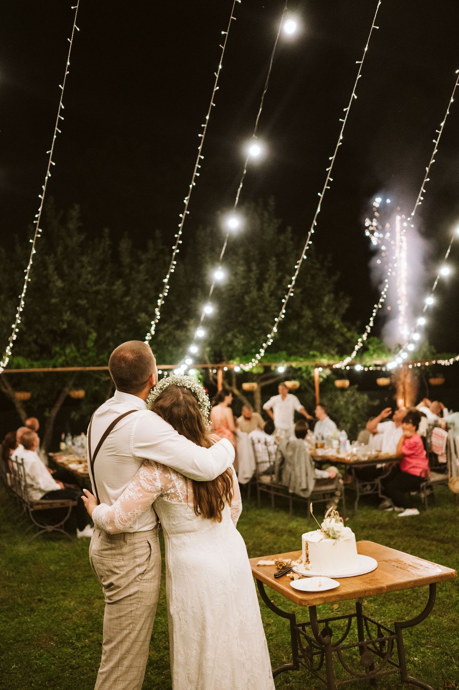 Couple embraces while watching fireworks under string lights at outdoor wedding reception.