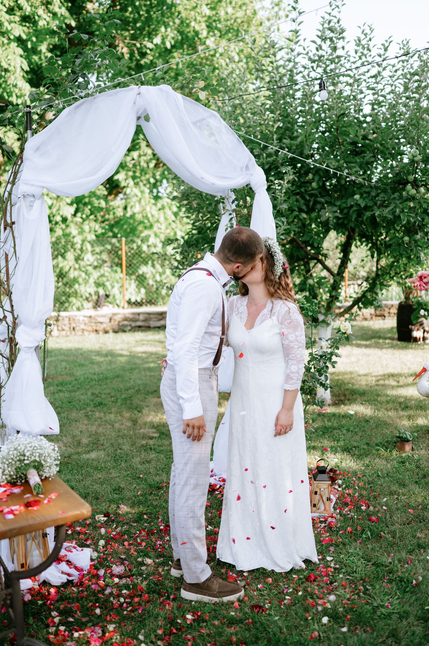 Couple in white attire standing beneath a draped wedding arch with rose petals on the ground.