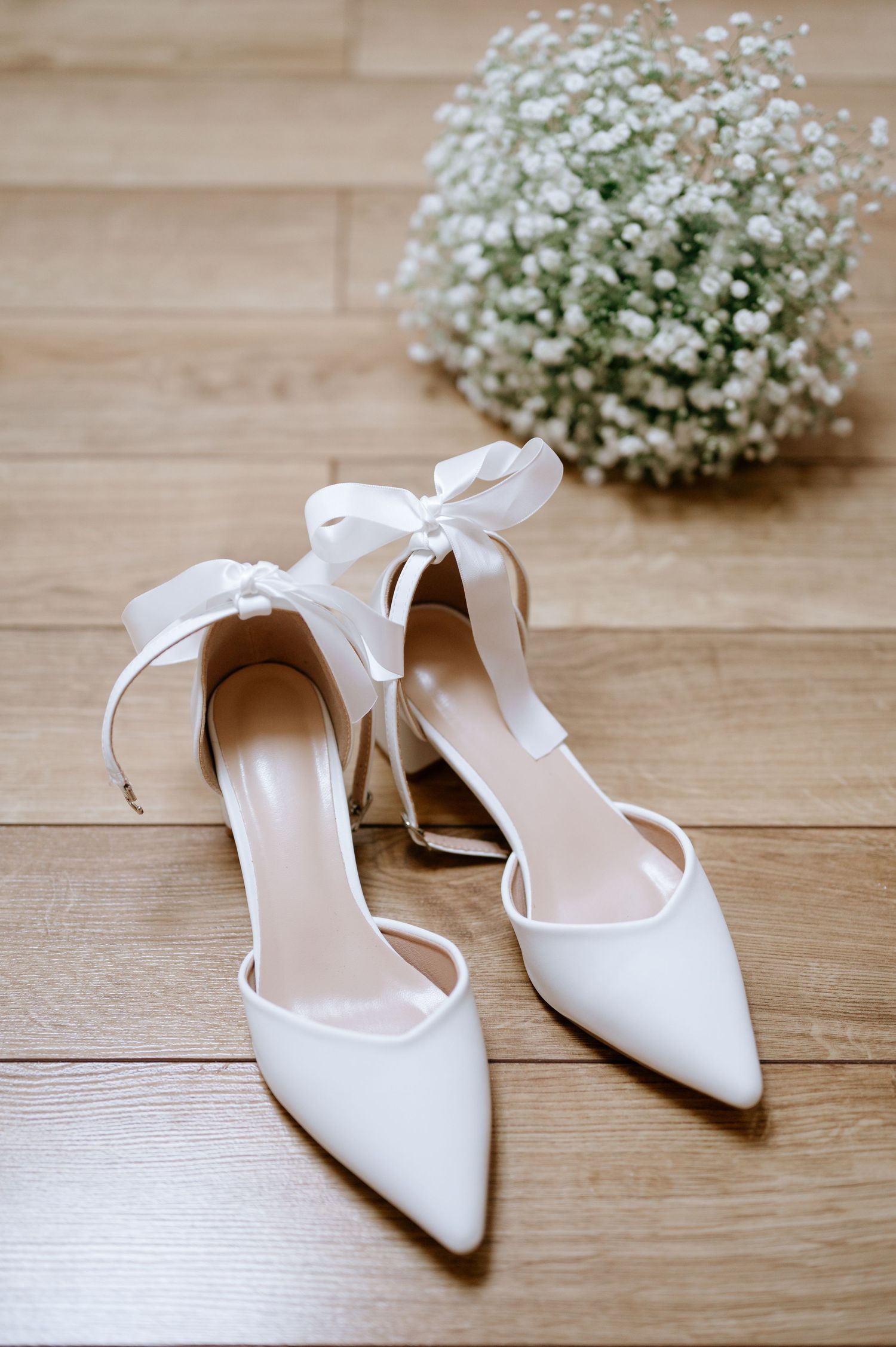 Elegant white bridal shoes with bow details and baby's breath bouquet on wooden floor.