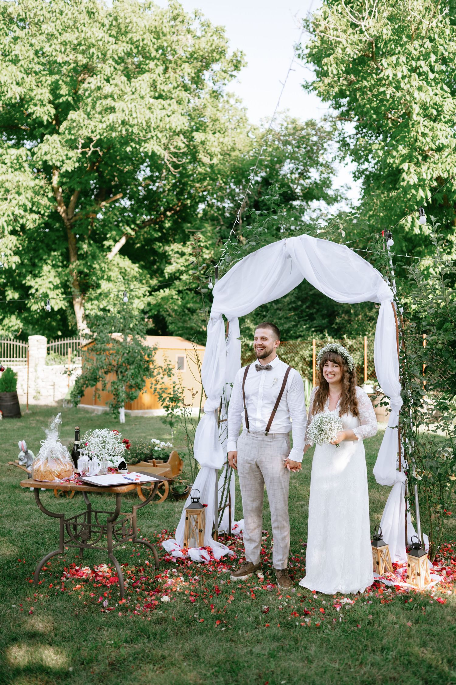 Garden wedding ceremony space decorated with white fabric arch and vintage furniture under trees.