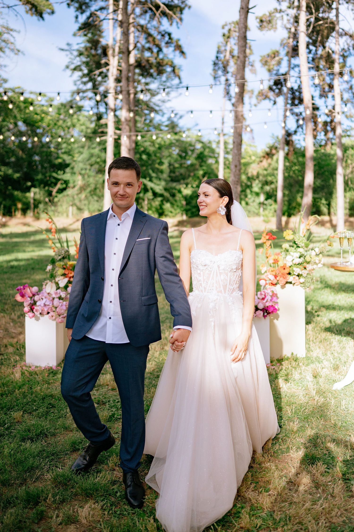 Wedding couple poses in a forest setting with string lights and floral arrangements on white pedestals.
