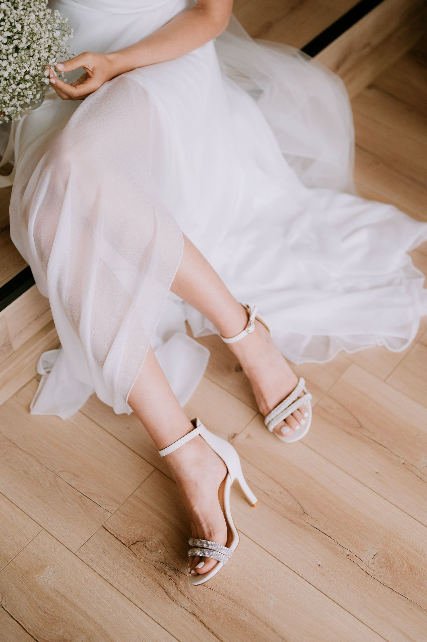 Close-up of white bridal shoes and dress hem on light wooden flooring.