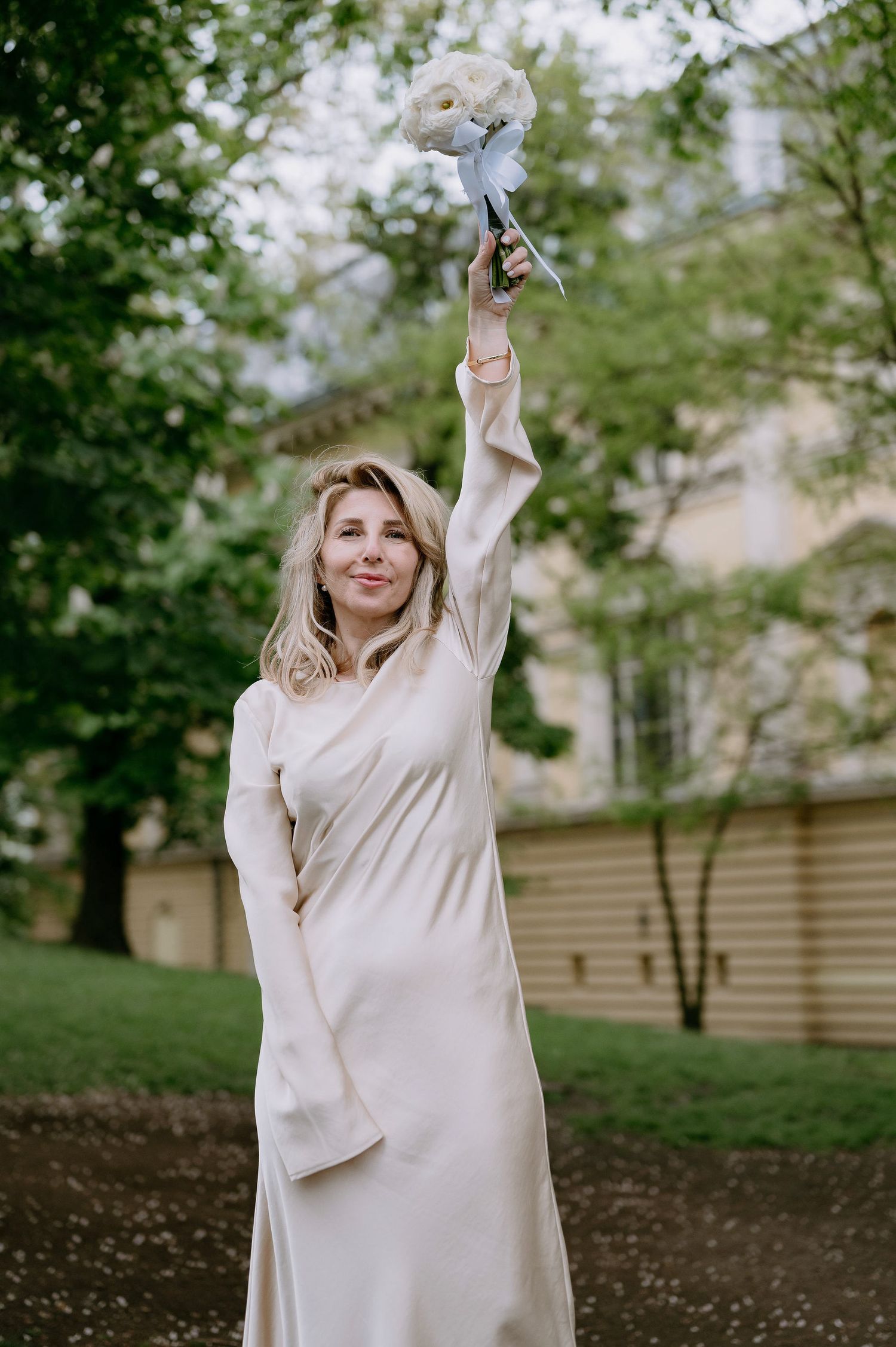 A person in a flowing cream dress holds a bouquet high in the air against trees.