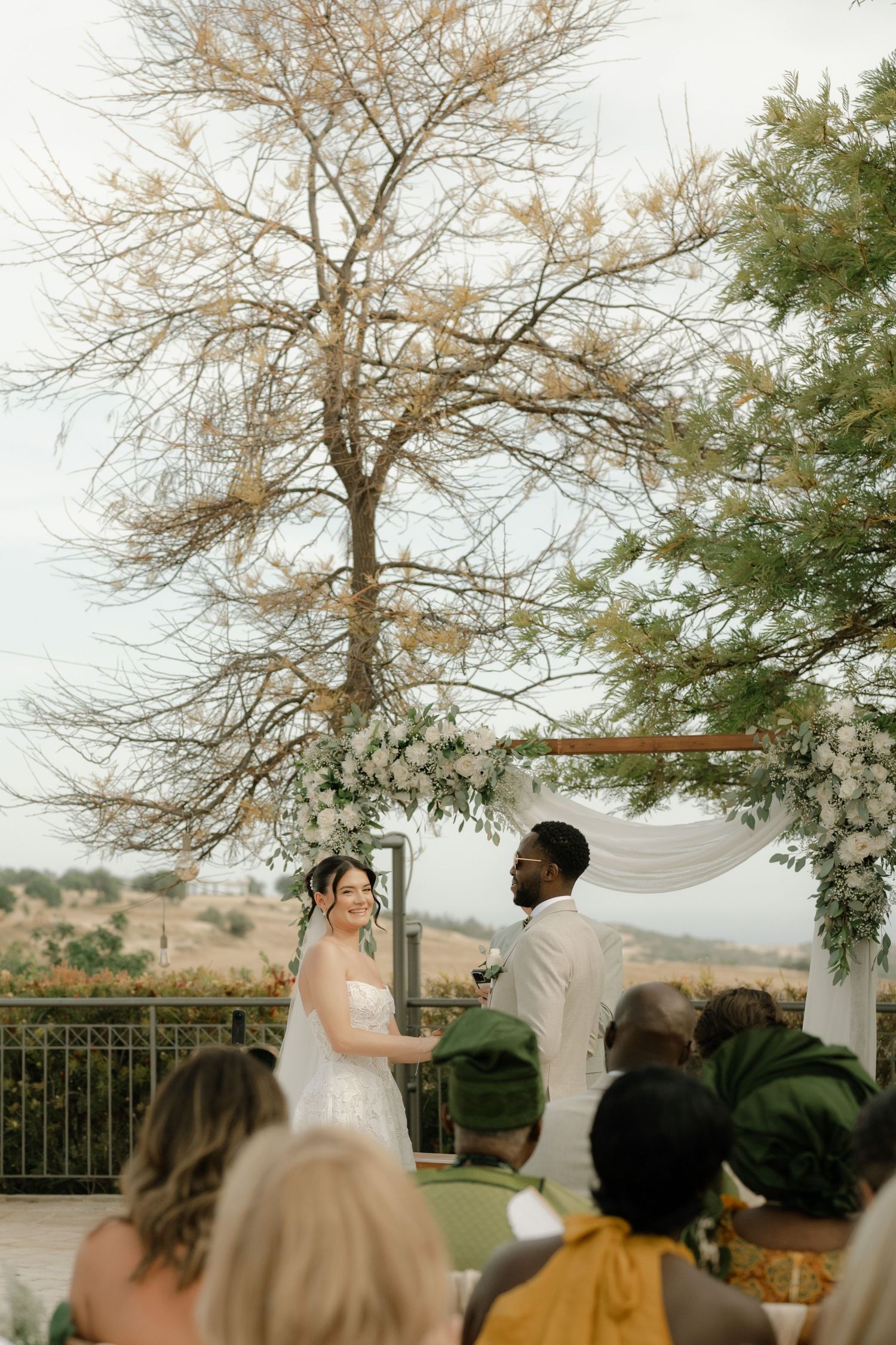 Outdoor wedding ceremony with bride in white gown and groom in ivory suit exchanging vows under floral arch.