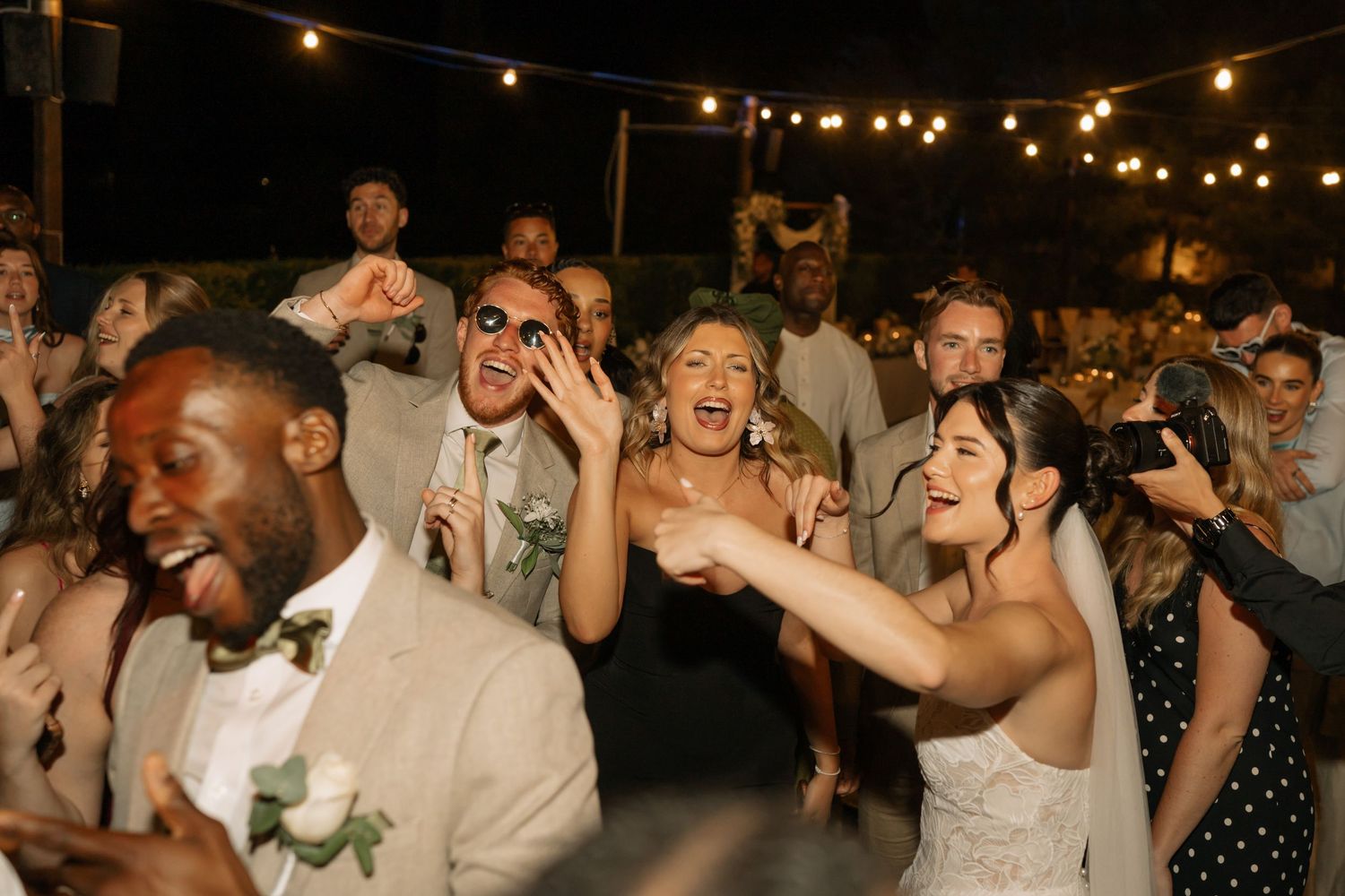 Joyful wedding guests dancing and celebrating at an outdoor nighttime reception with string lights.
