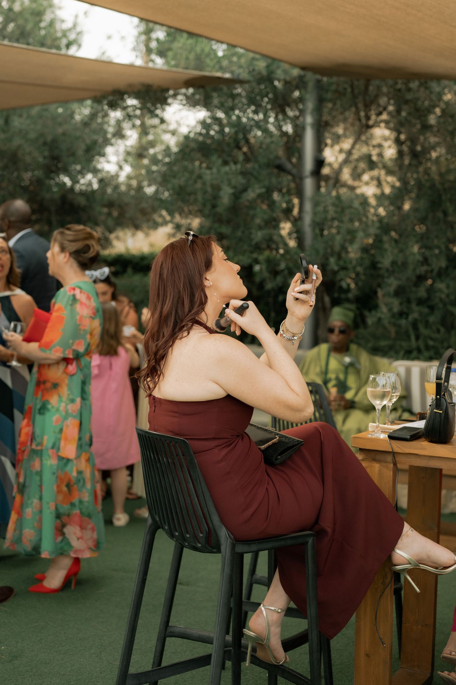 Woman in burgundy dress sits at outdoor bar, laughing while holding phone at elegant garden party.