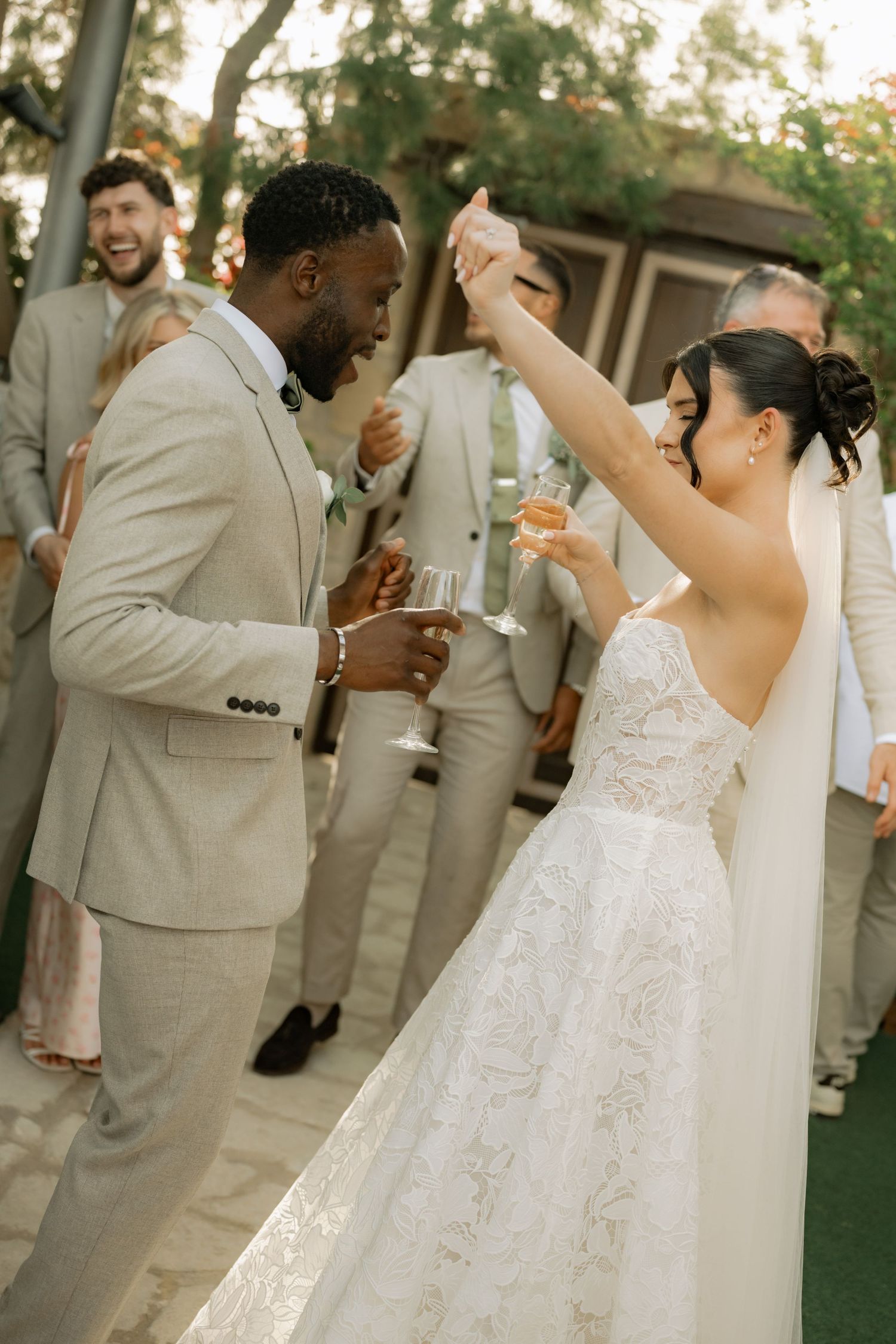 Bride and groom dance joyfully outdoors, raising champagne glasses at their wedding reception.