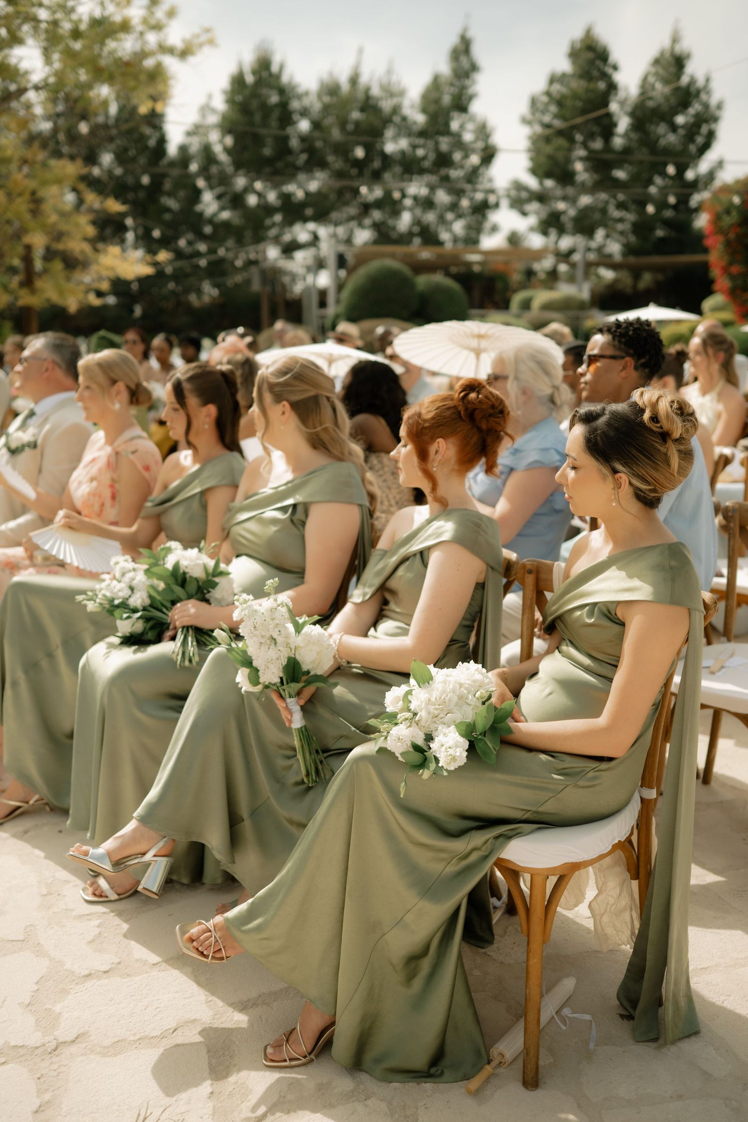 Bridesmaids in sage green satin dresses holding white bouquets seated at an outdoor wedding ceremony.