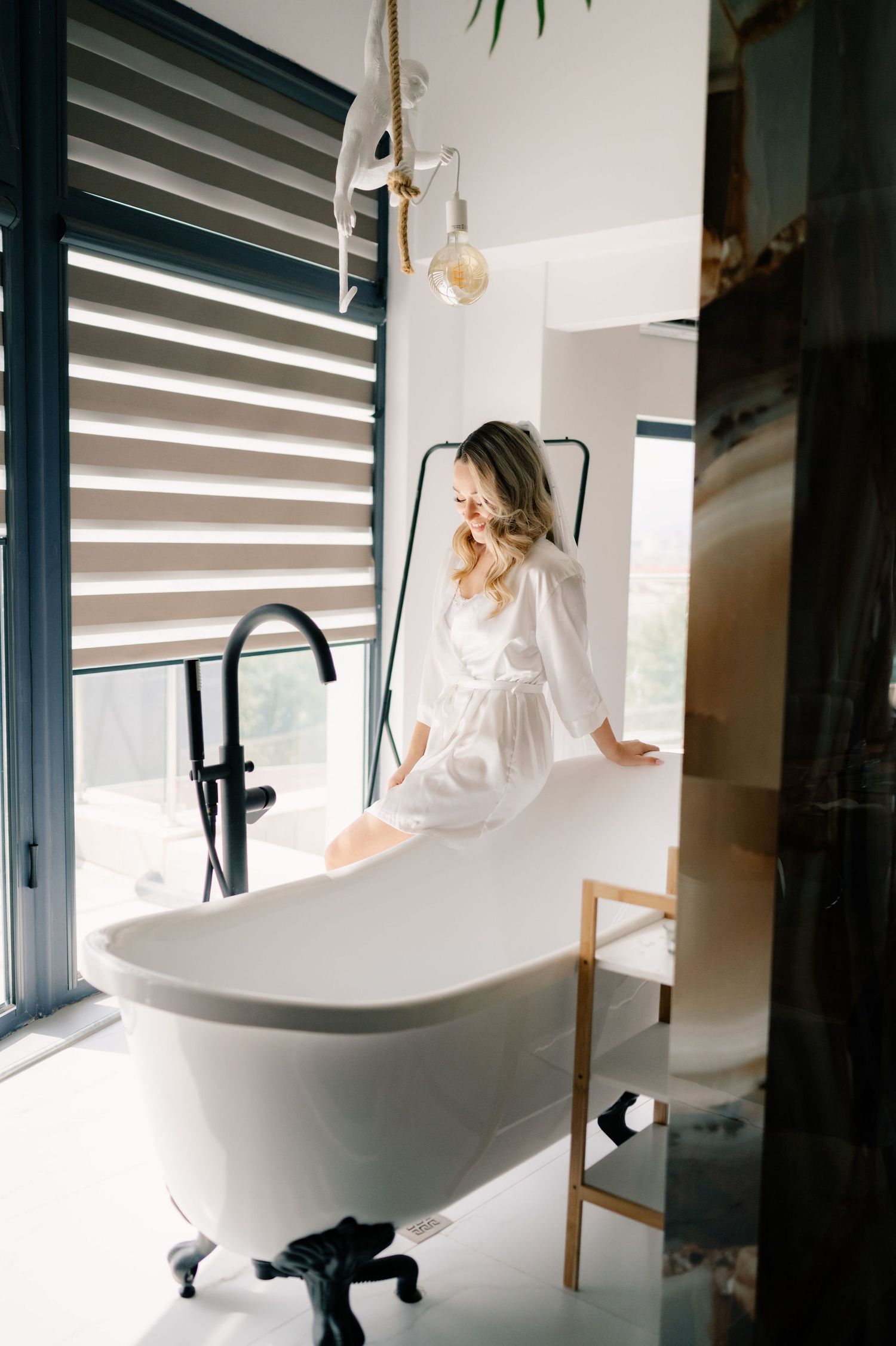 A white freestanding bathtub with black fixtures sits near a window with modern blinds in a luxurious bathroom setting.