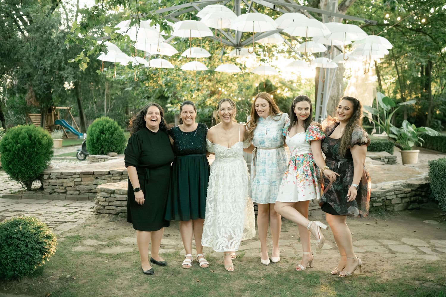 Group of friends in formal attire pose together at an outdoor garden party with white umbrellas in the background.