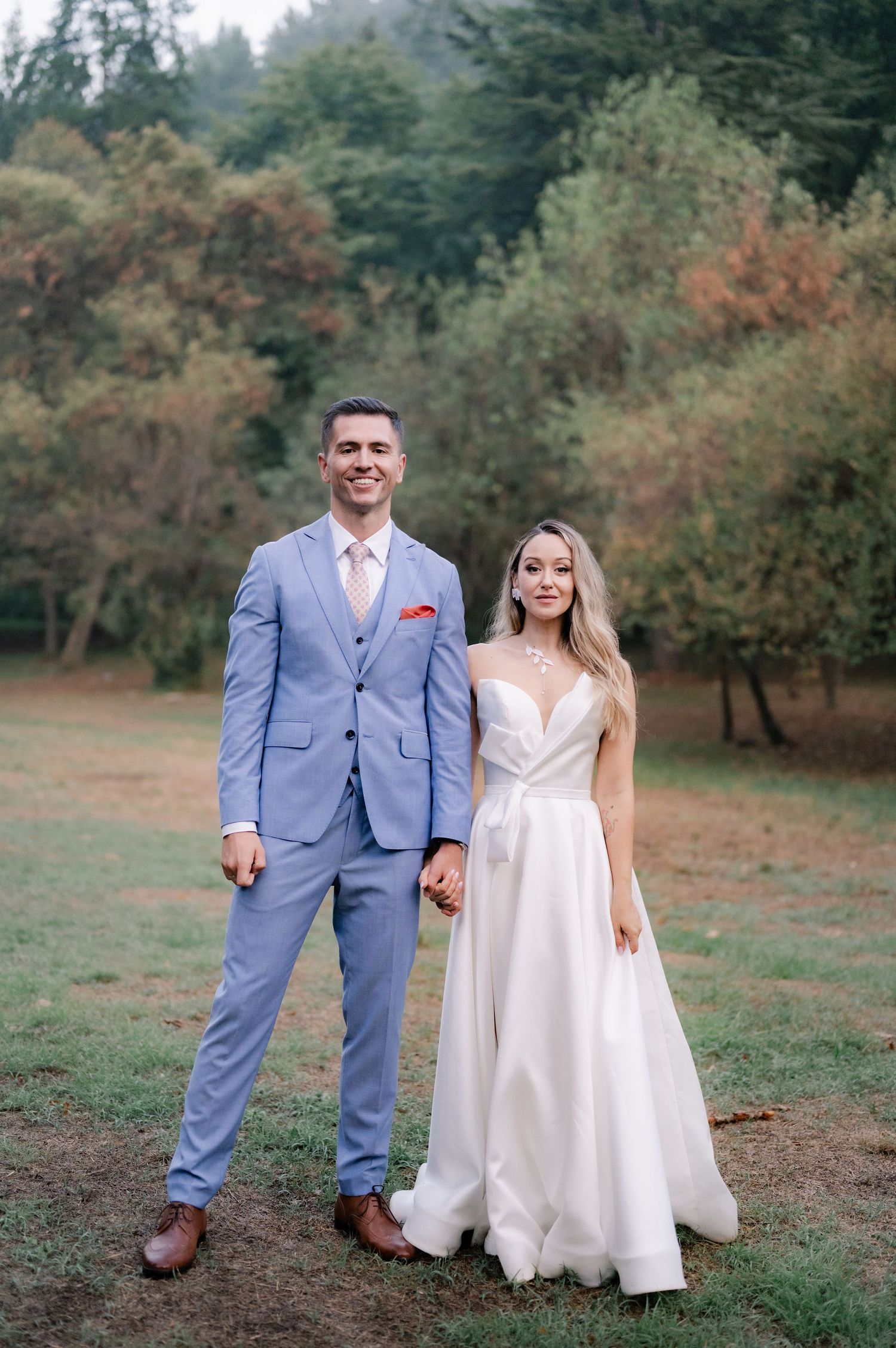 A couple in wedding attire poses together in a scenic outdoor setting with autumn trees in the background.