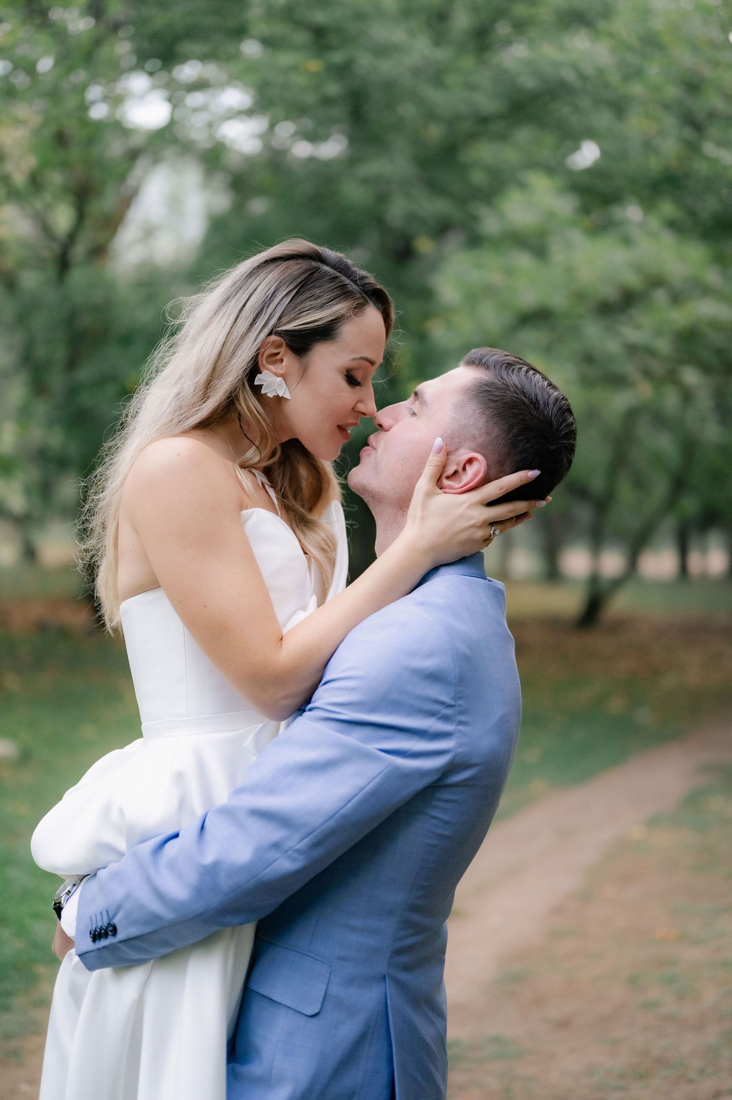 A couple in wedding attire share a romantic kiss in a park setting with green trees in the background.