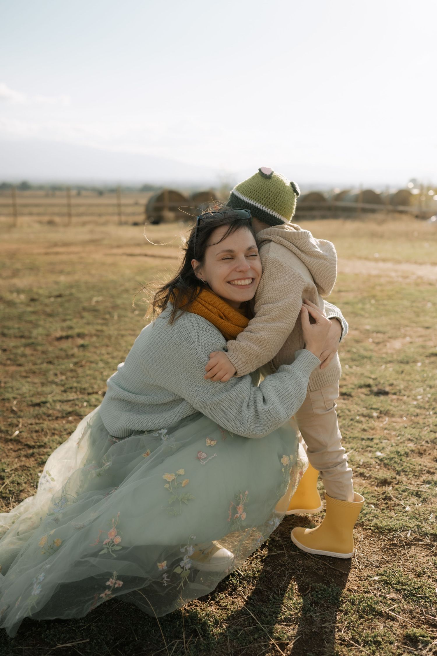A tender embrace between parent and child in a field, wearing casual clothing and yellow boots on a cloudy day.