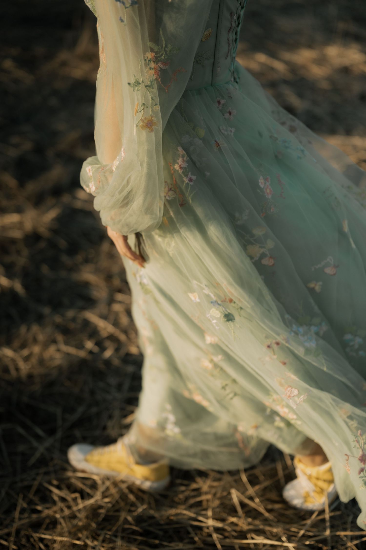 A flowing mint green dress with floral embroidery paired with yellow sneakers creates a whimsical contrast on natural ground.