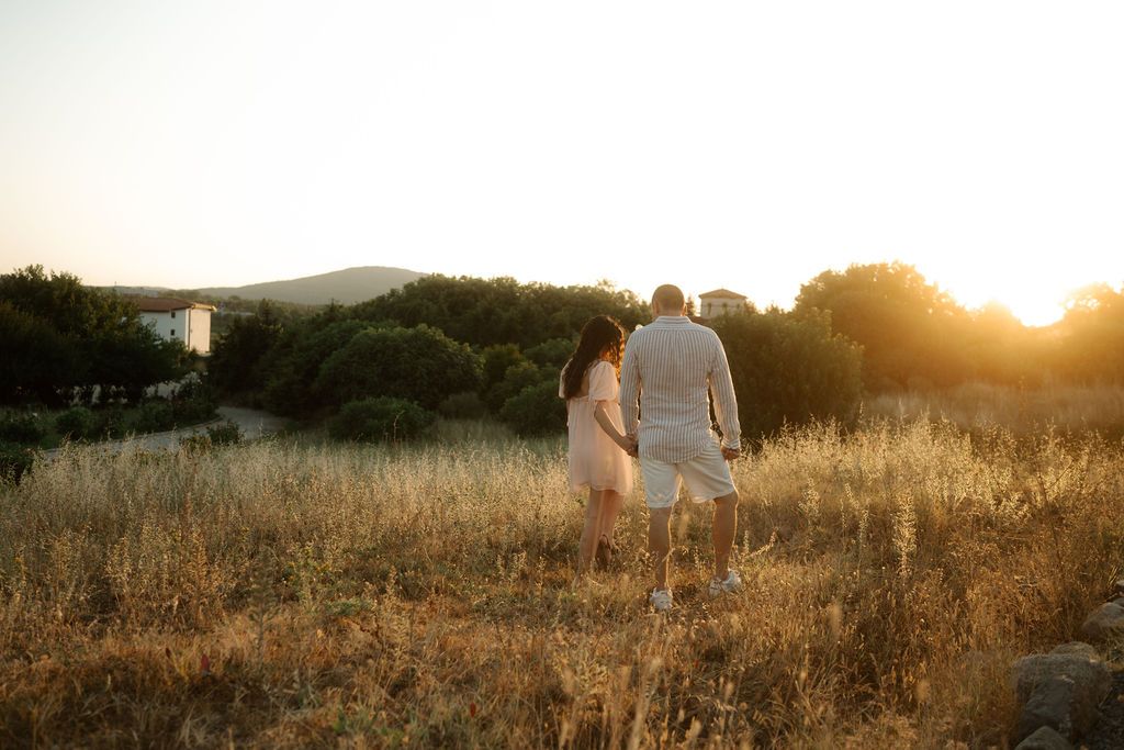A couple walks through a golden field at sunset holding hands while silhouetted against the warm glowing sky.