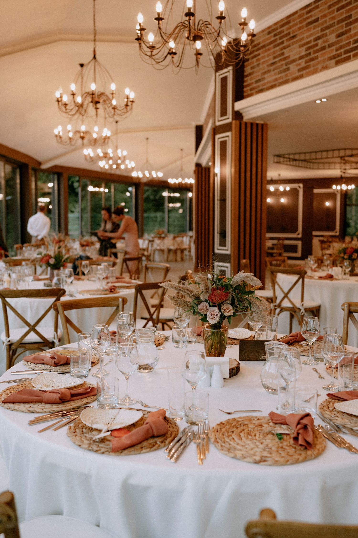 Elegant wedding reception tables with white linens, chandeliers, brick walls, and peach napkins in rustic venue setting.