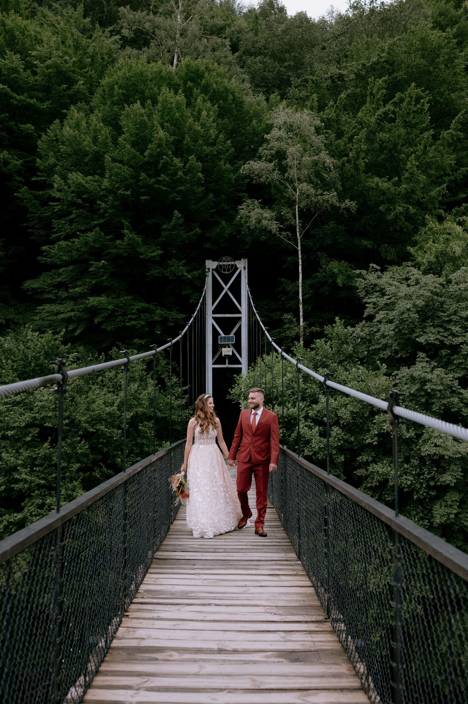 A couple walks together across a scenic wooden suspension bridge surrounded by lush green forest.