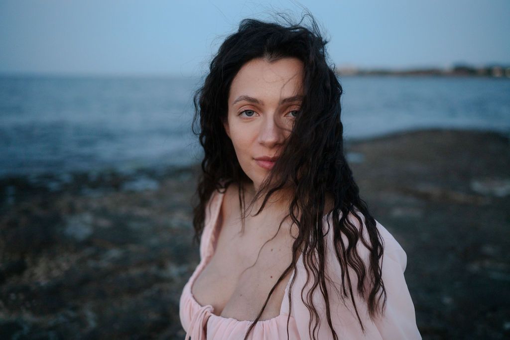 A person with dark wavy hair in a pink top poses near the ocean at dusk.