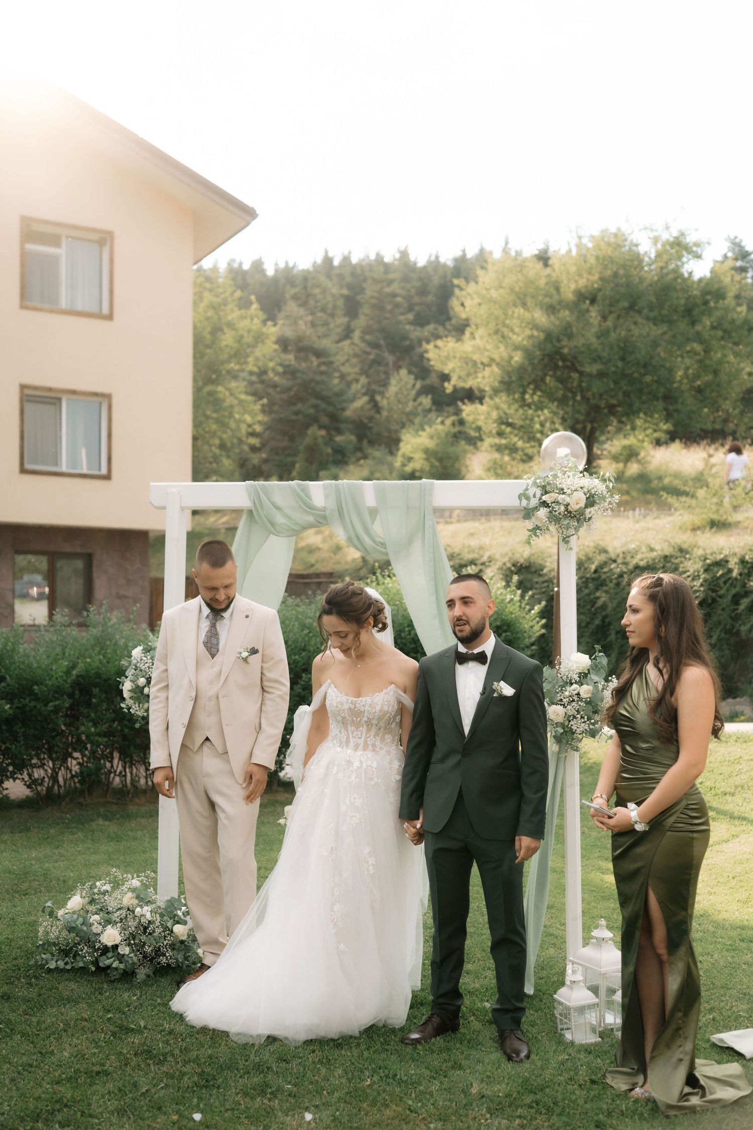 A wedding party stands together outdoors in a garden setting with white decorative panels and greenery in the background.