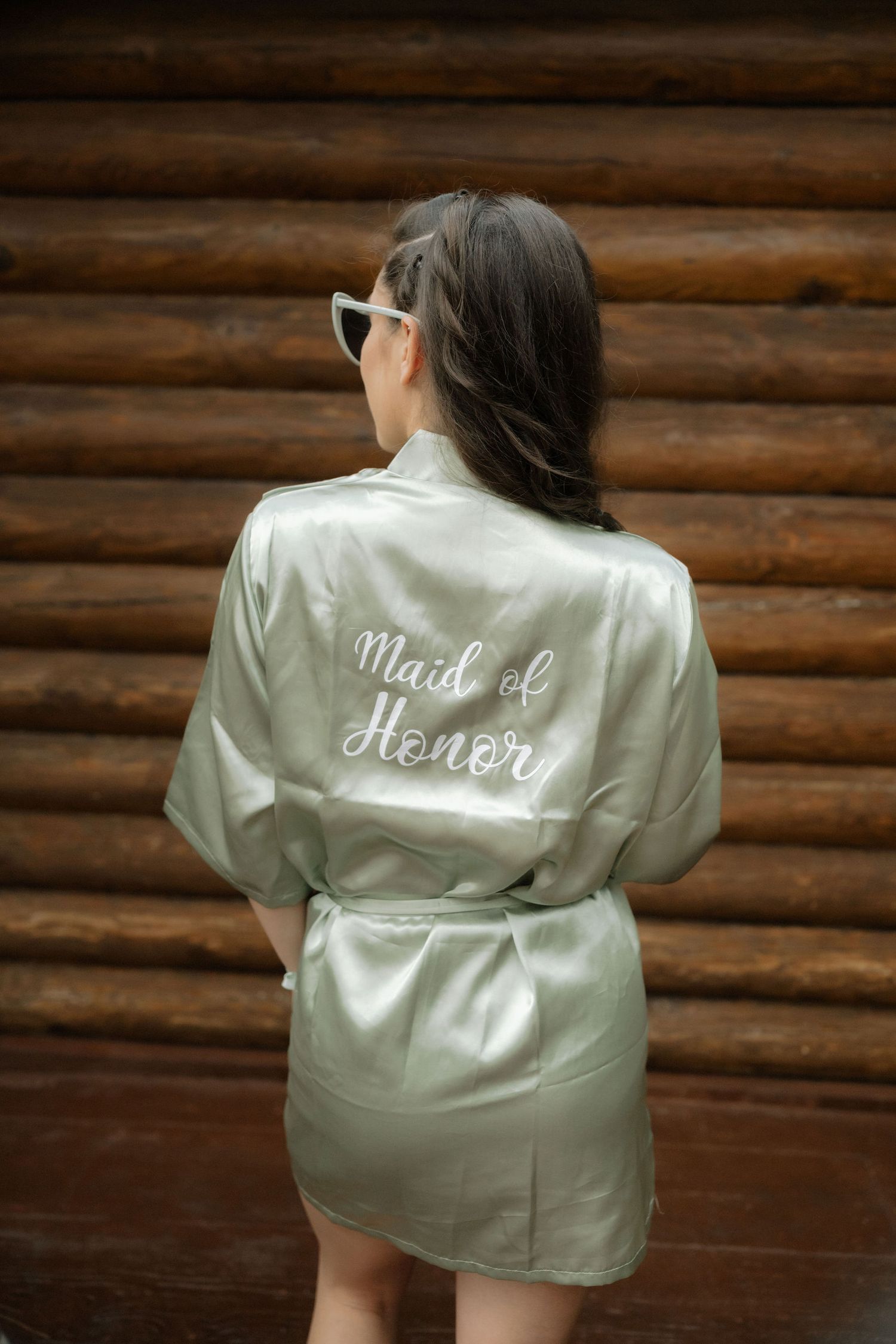 A woman wearing a mint green satin robe with 'Wifey' embroidered text on the back against a rustic log cabin wall.