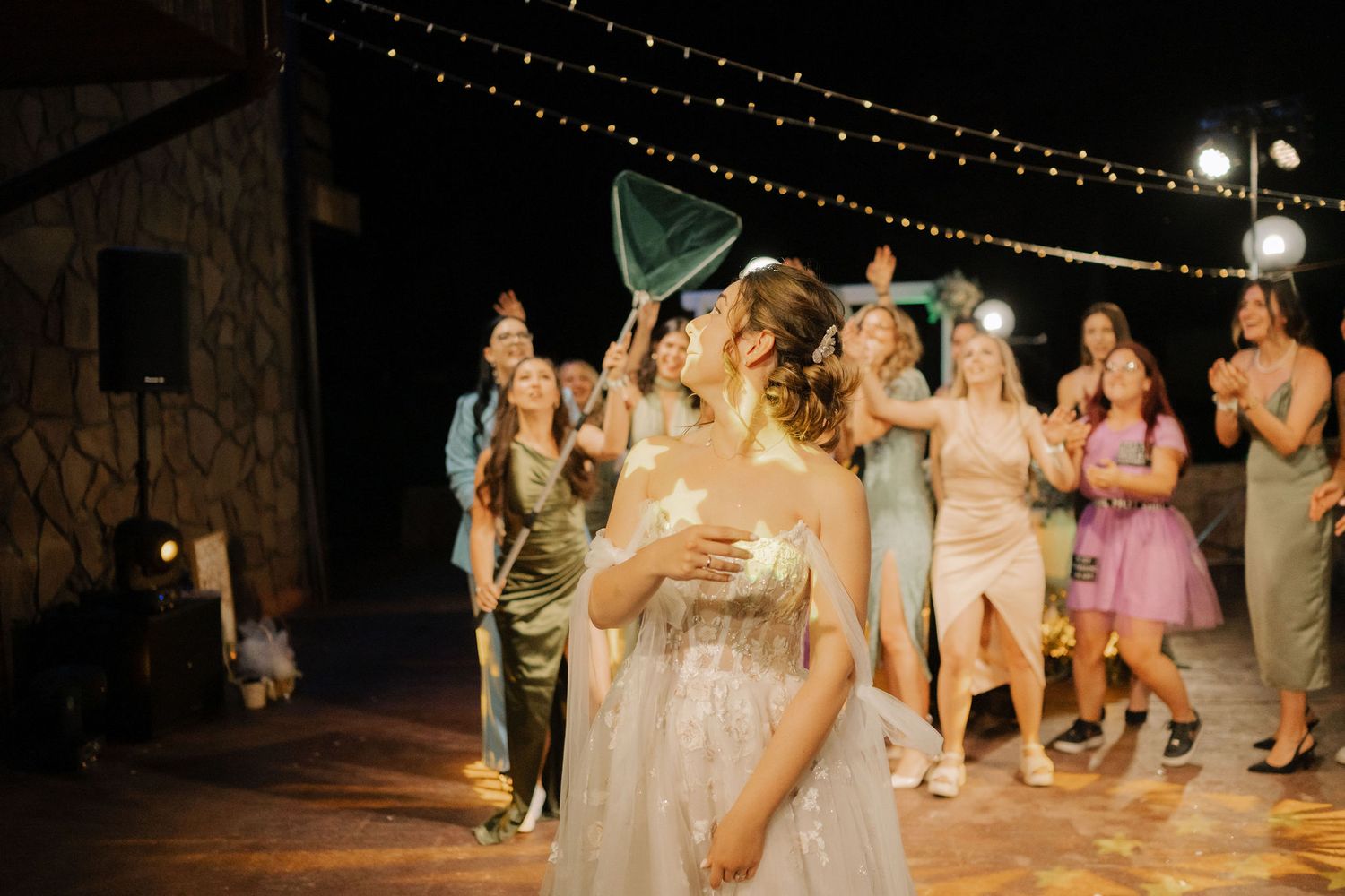 Celebratory dancing scene at an outdoor evening party with string lights and stone wall backdrop.