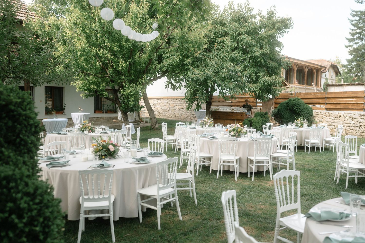 Elegant outdoor wedding reception setup with white chairs and tables arranged on a lush green lawn under string lights.