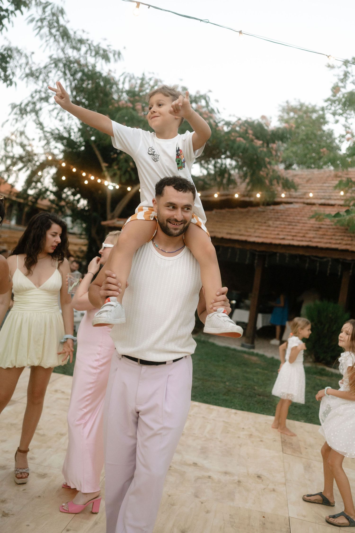 People dancing and celebrating at an outdoor party with string lights and rustic decor in the background.