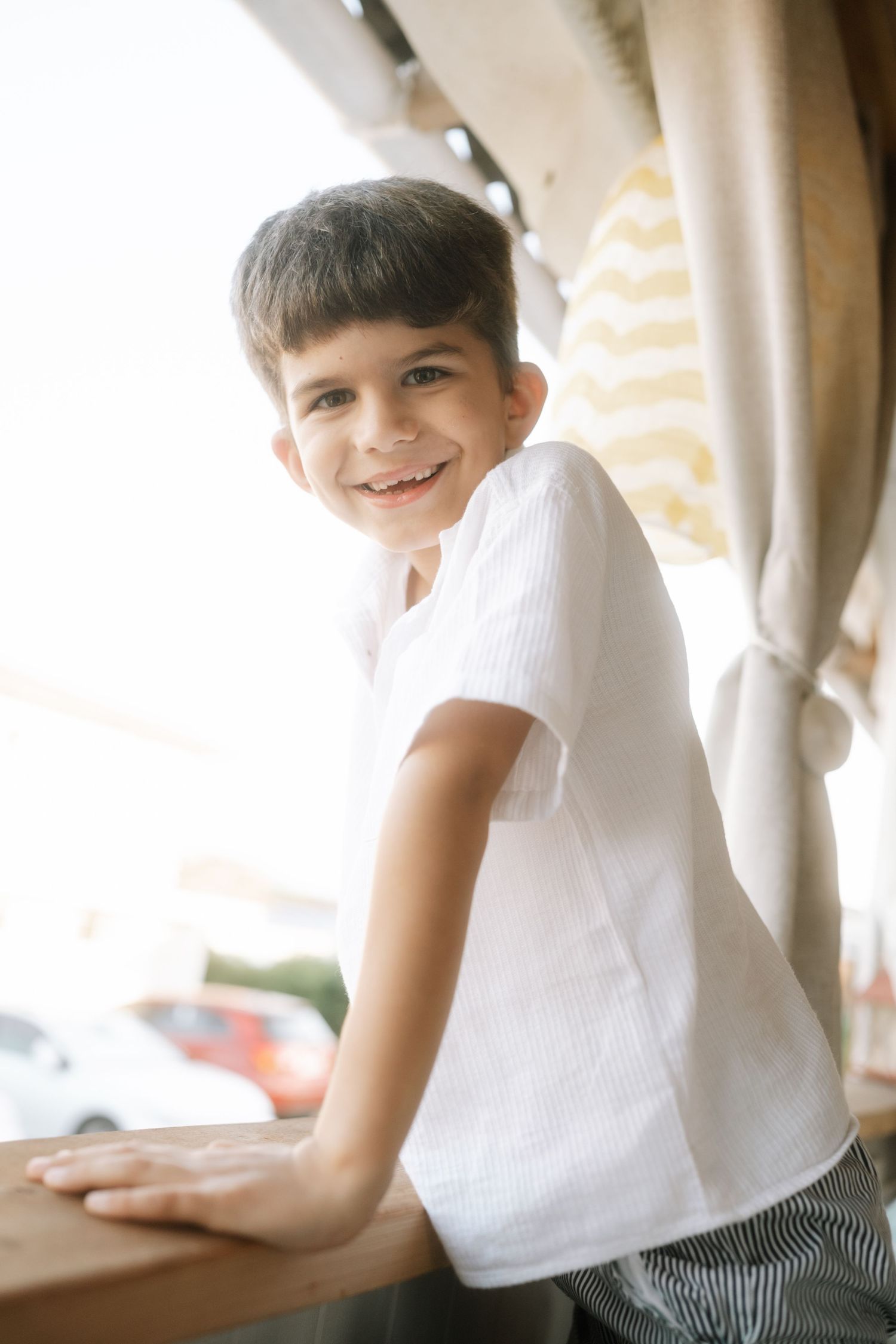 A child in a white t-shirt leans against a ledge by a window with curtains in a casual indoor setting.