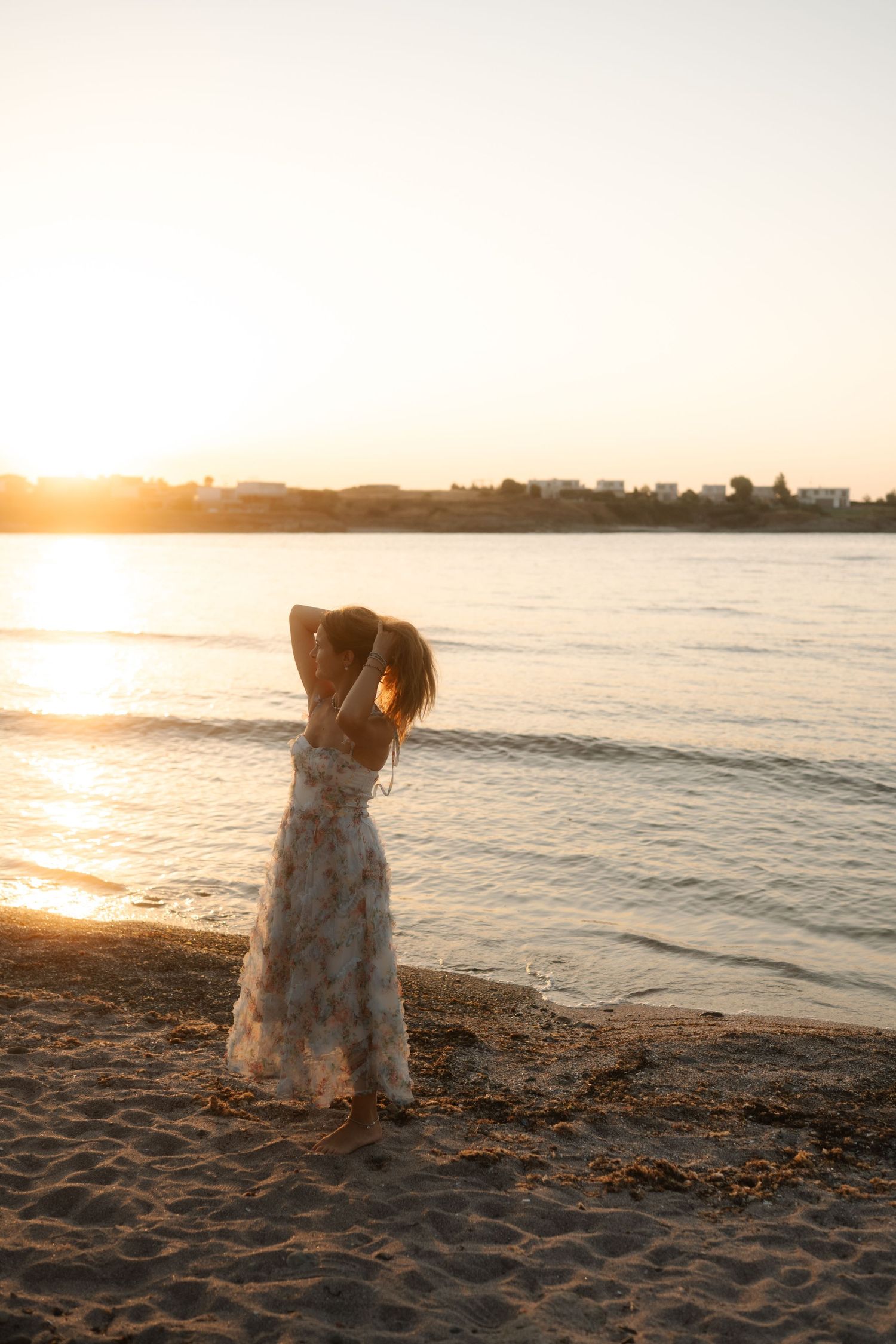 Figure in a white dress sits on sandy beach watching golden sunset over calm waters.