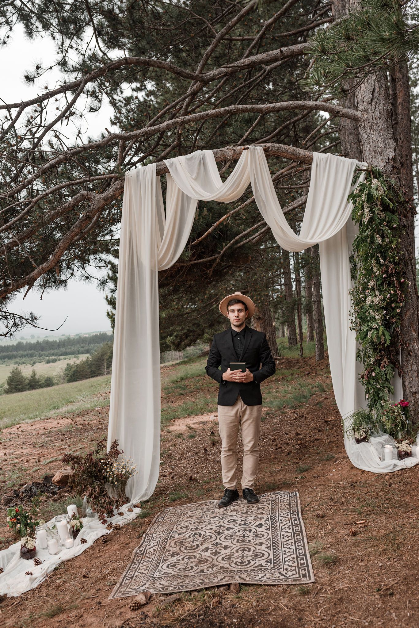 Elegant white fabric arch and vintage rug create a romantic woodland ceremony setting among evergreen trees.