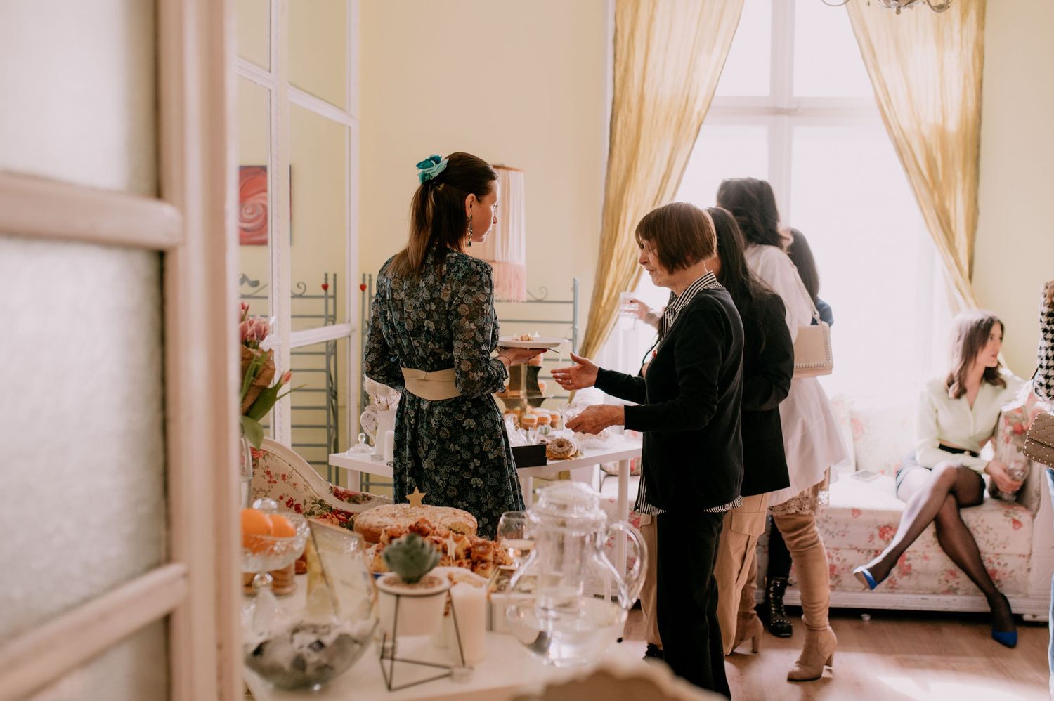 People gather in a bright room with soft curtains and decor for what appears to be a social event or celebration.