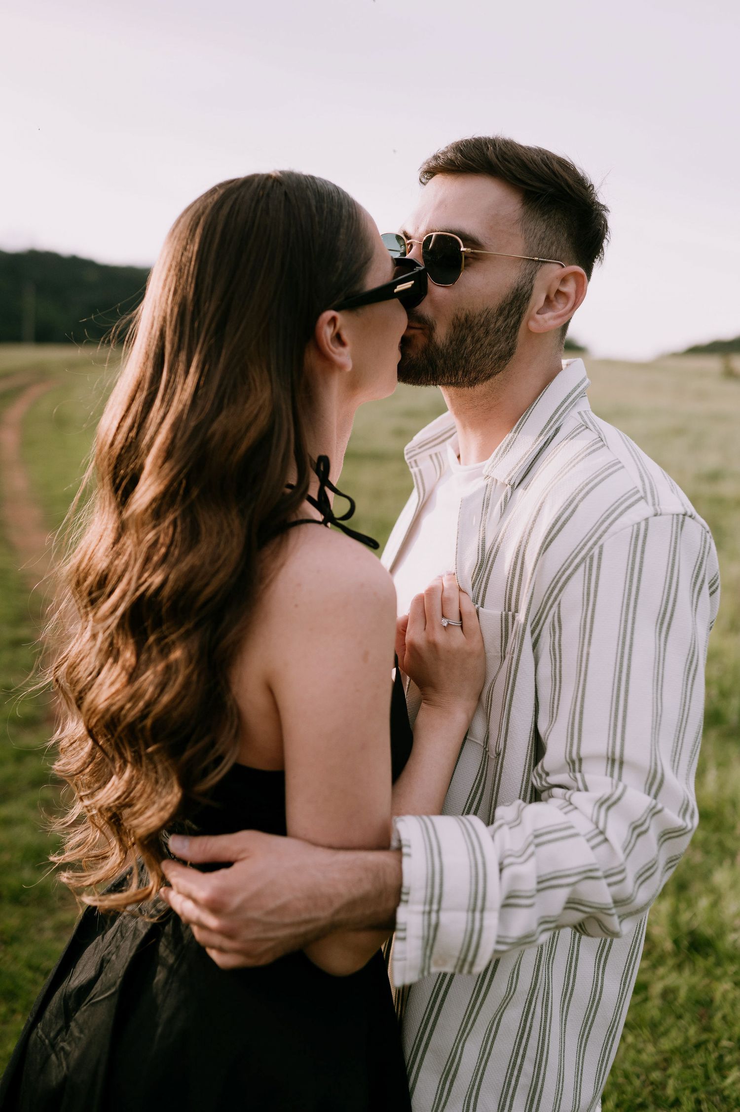 Romantic couple embracing in a grassy field at sunset, with long wavy hair and striped shirt details.