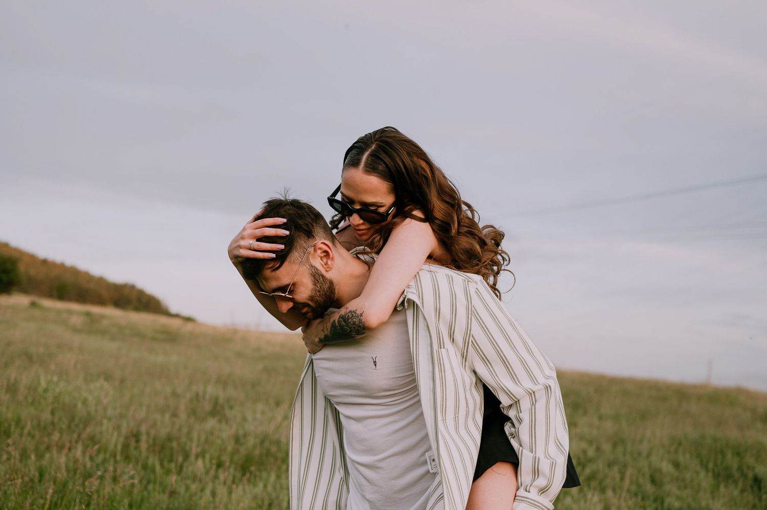 A couple embraces lovingly in a field of tall grass under an overcast sky during an outdoor photoshoot.