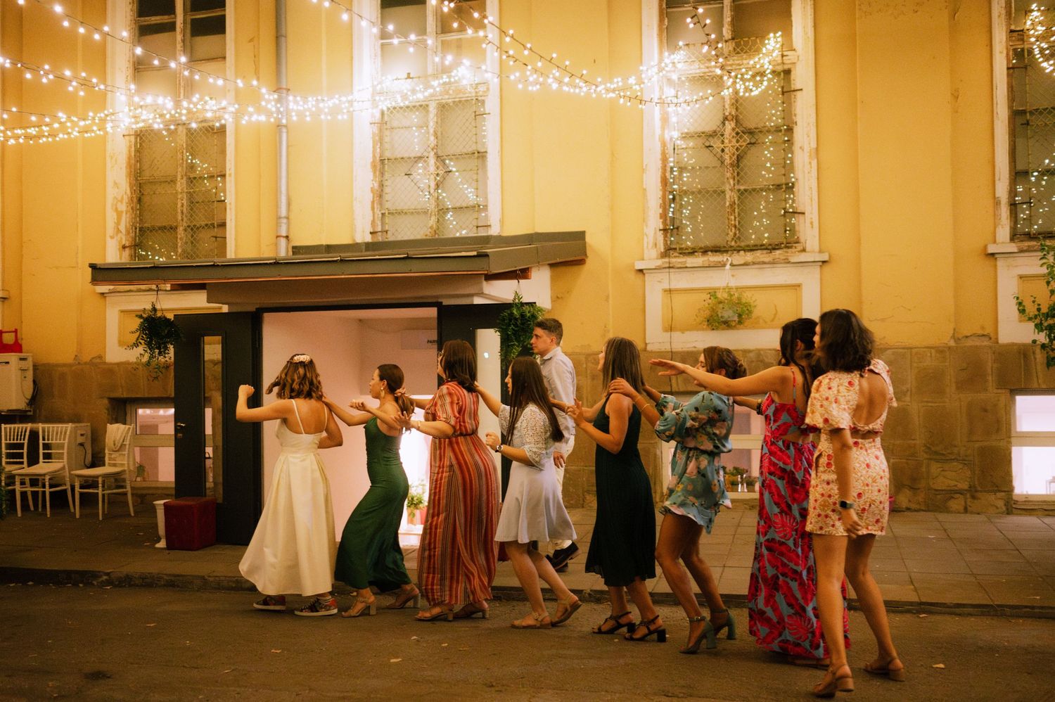 Wedding guests dancing and celebrating at an outdoor evening reception with glowing string lights and historic building facade.