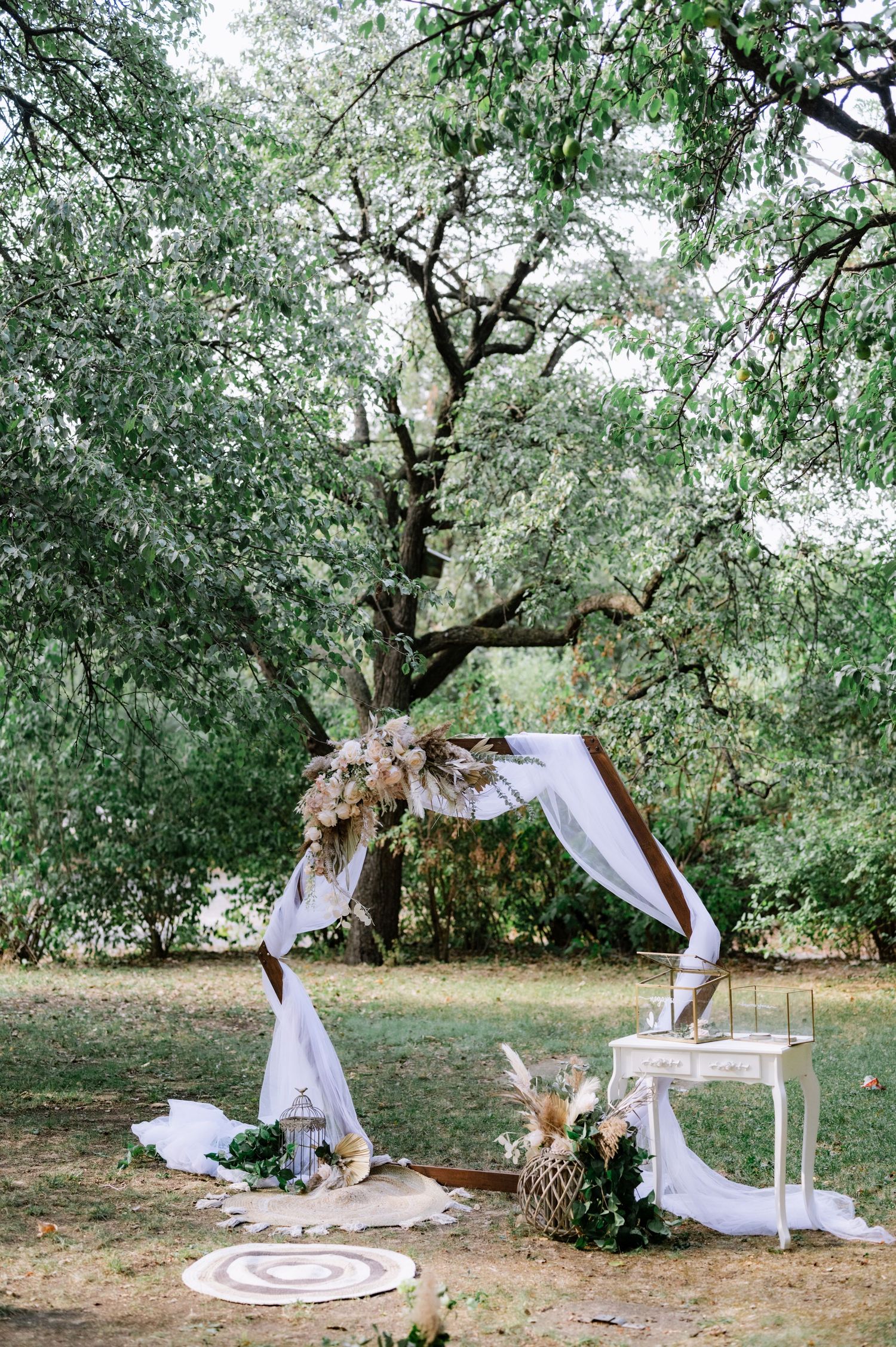 Romantic outdoor picnic setup with white draping, rugs, and cushions under a blooming tree in a garden setting.