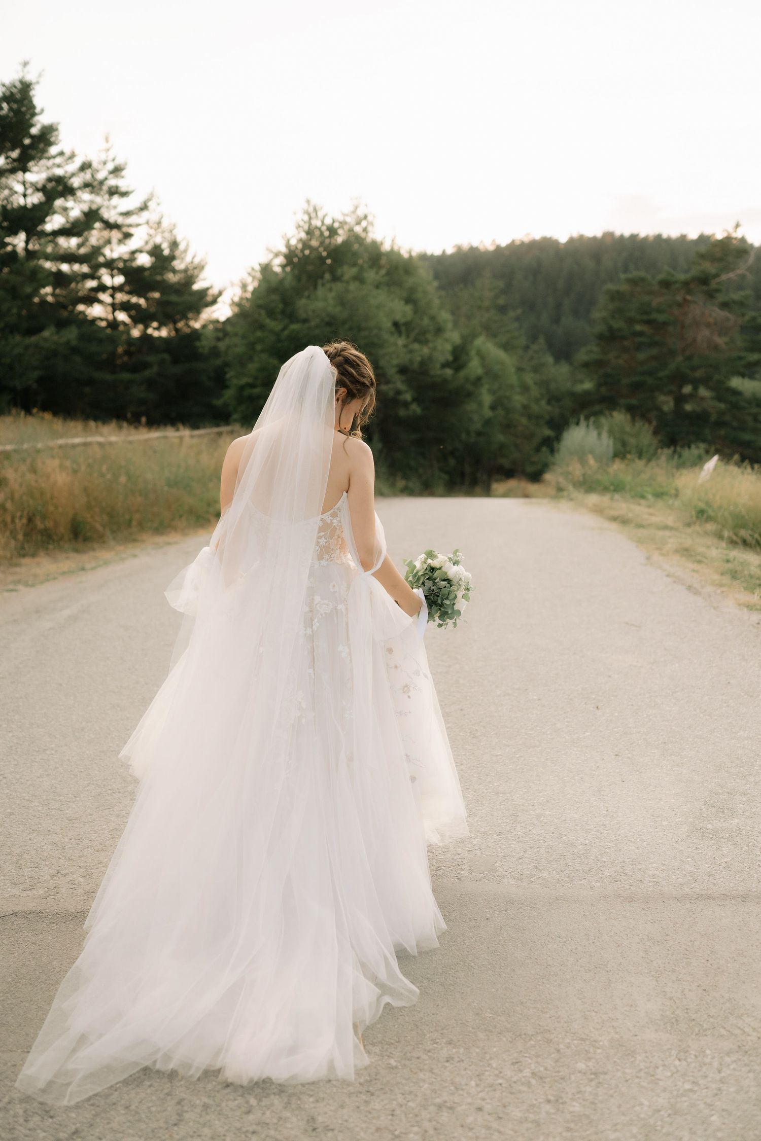 A figure in a flowing white wedding gown and veil walks along a serene dirt path surrounded by pine trees.