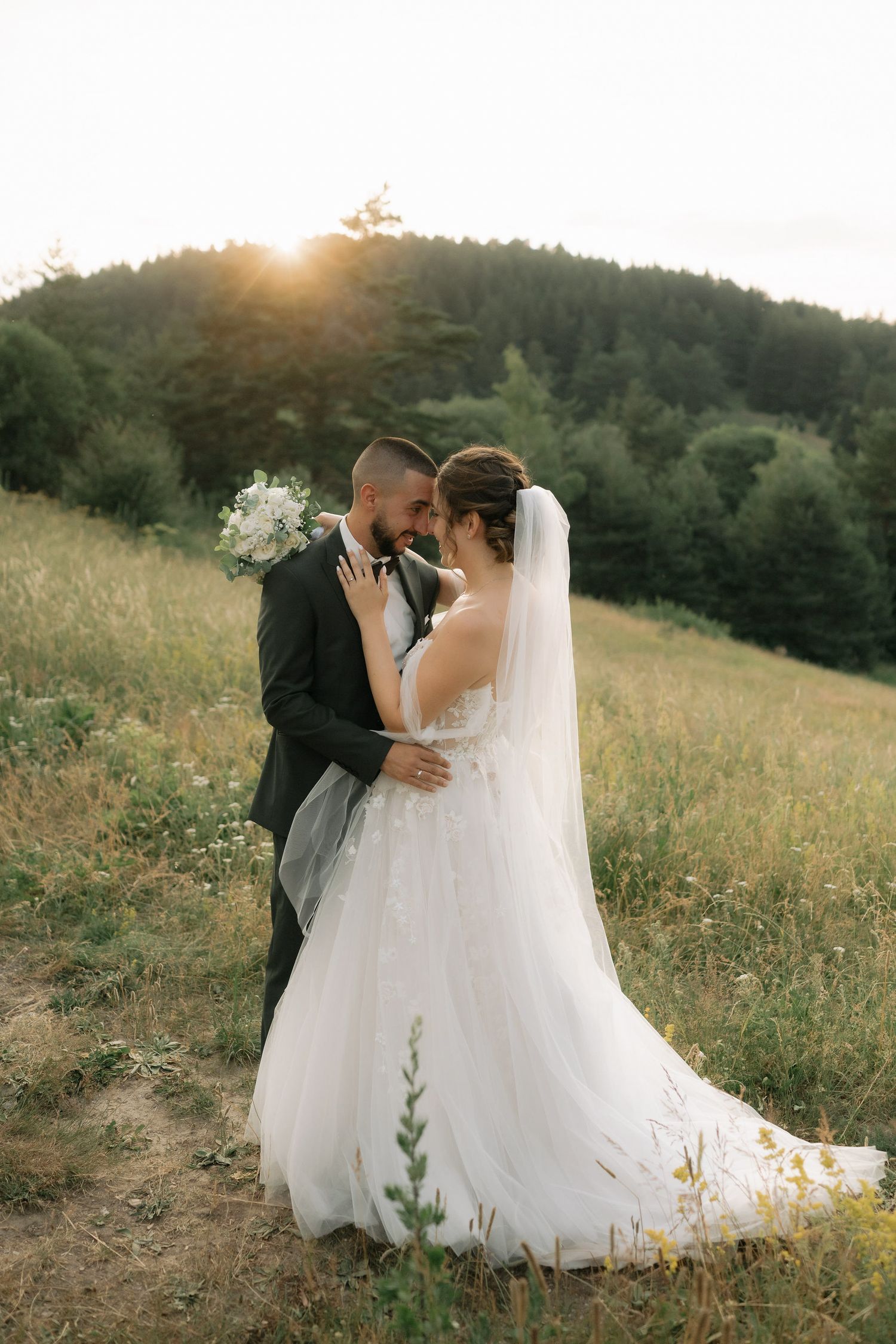 A romantic wedding couple embraces in a sunlit meadow at golden hour with flowing white dress and mountain backdrop.