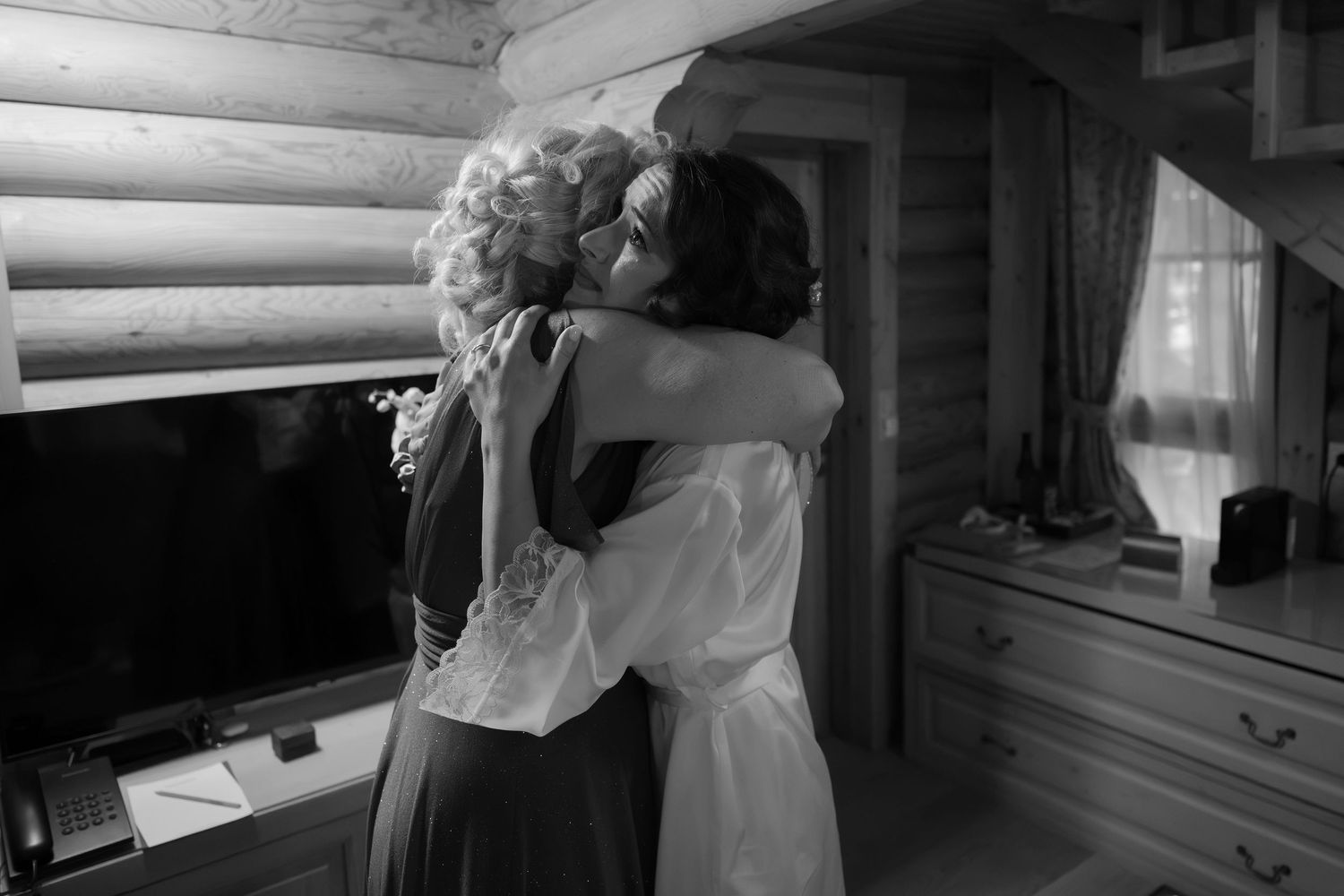 Black and white photo of two women sharing a warm embrace in a rustic log cabin room near a window.
