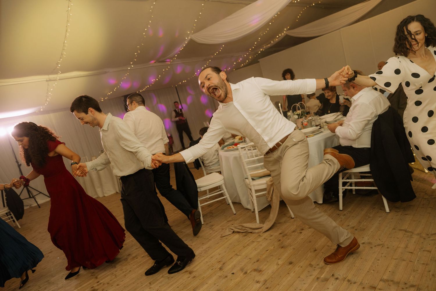 Guests dancing energetically at a wedding reception in a decorated tent venue with string lights.