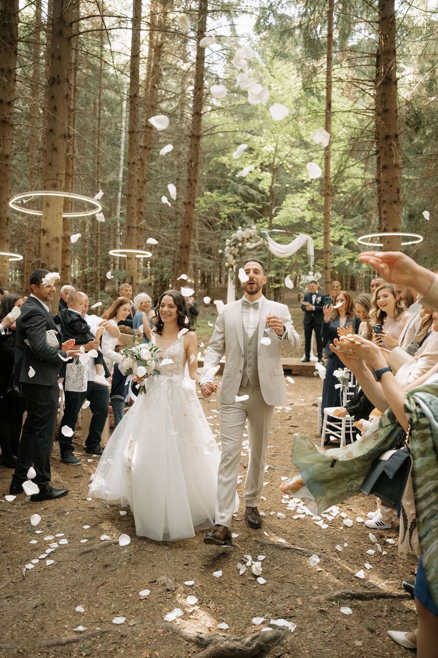 Bride and groom walk down forest aisle as guests toss white flower petals during outdoor woodland wedding ceremony.