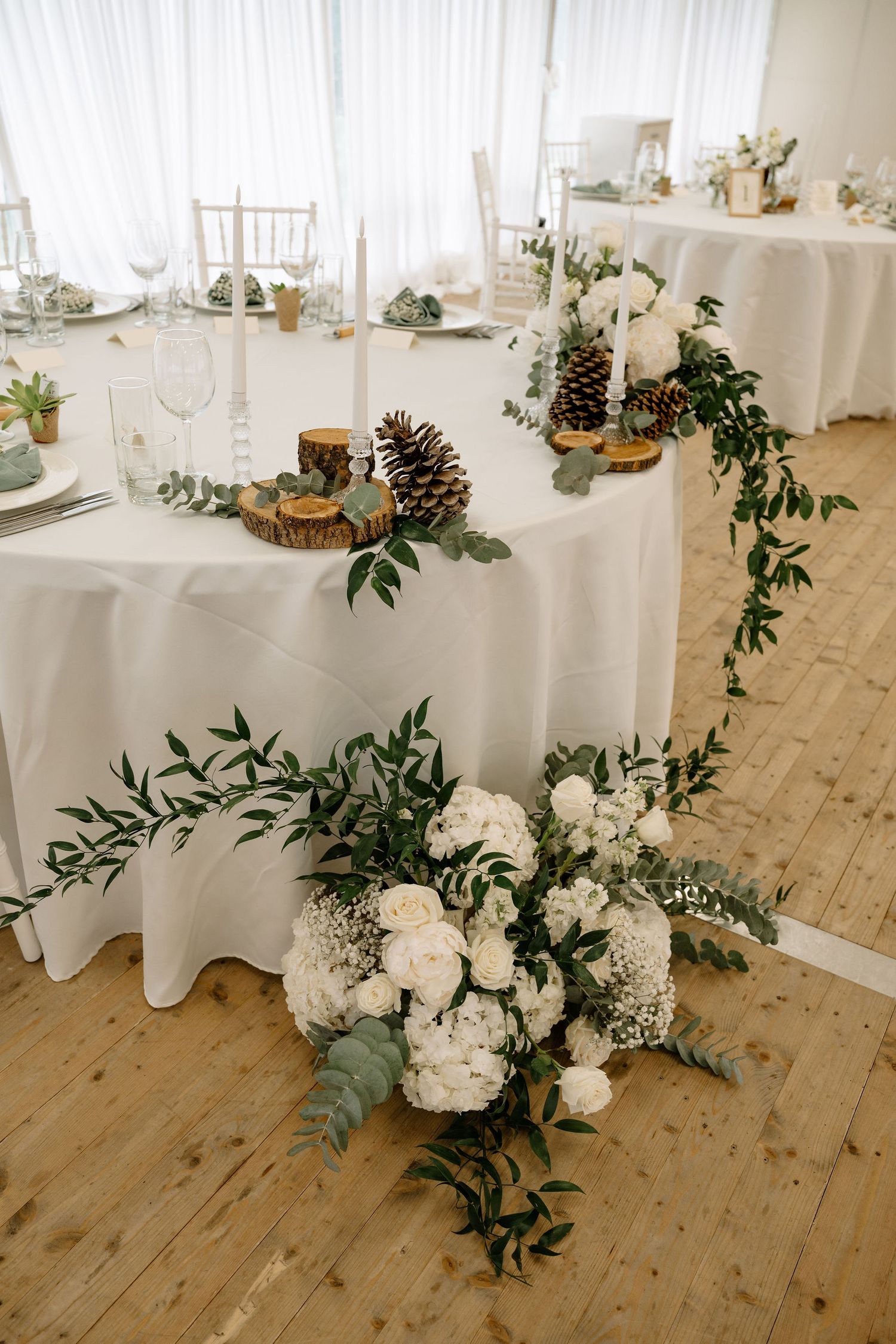 Elegant wedding reception table with white floral arrangements, greenery garland, pine cones, and candles on wood slice decor.