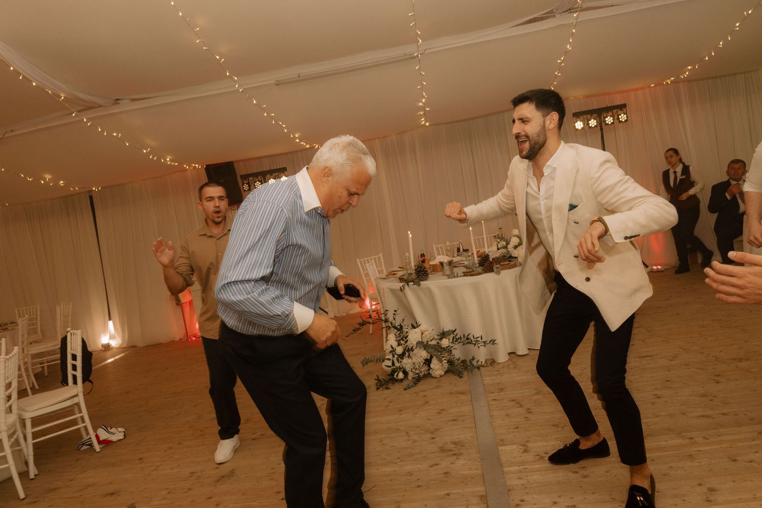 Two men dancing energetically at a wedding reception in a decorated venue with fairy lights.