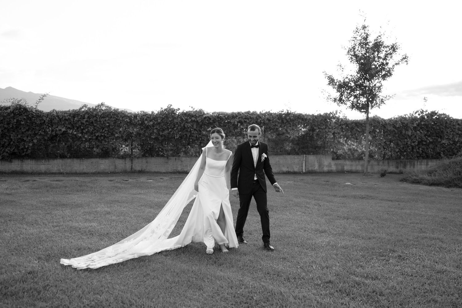 Black and white wedding photo of a couple walking together on a lawn with a long flowing bridal train.