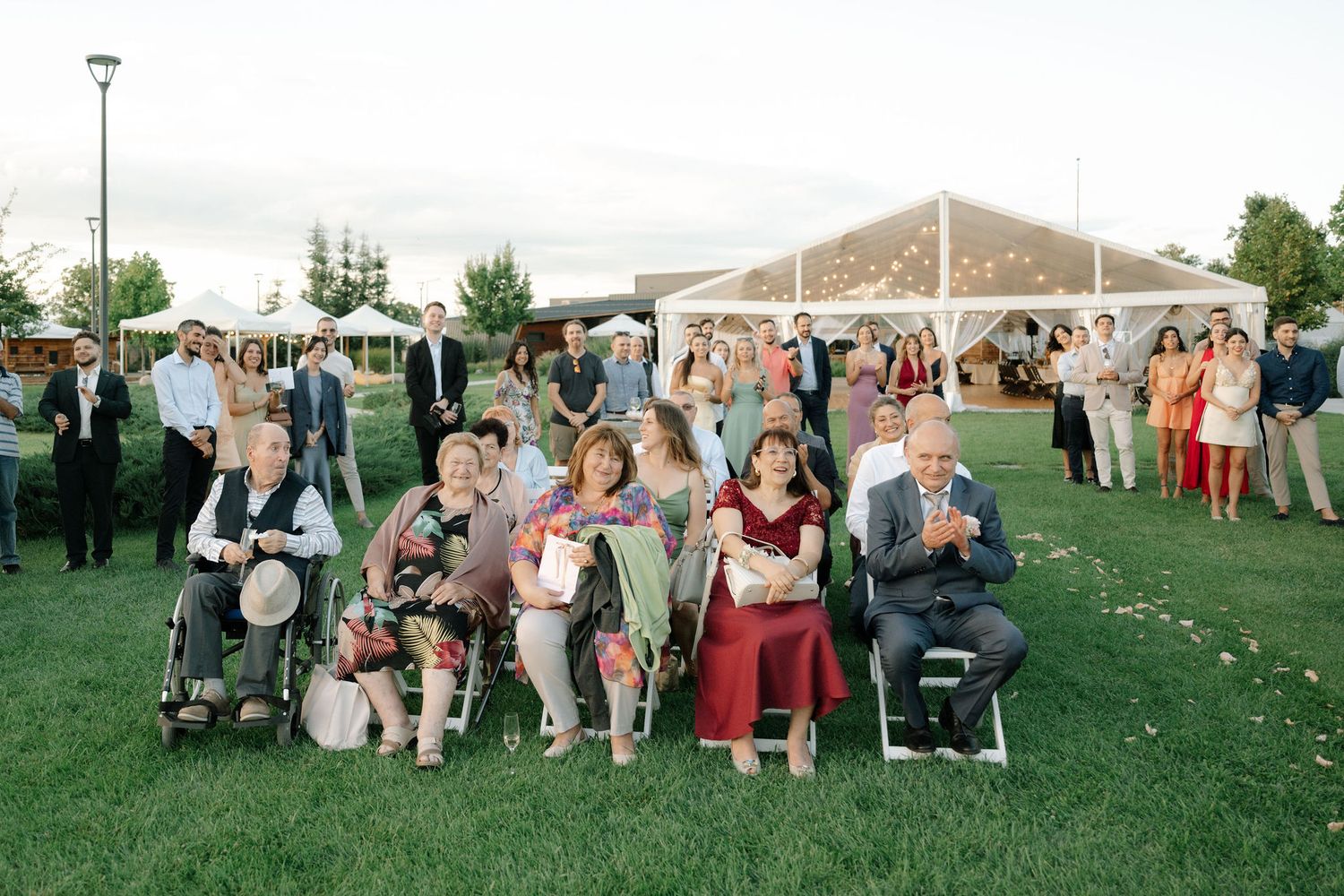 A panoramic view of wedding guests seated in white chairs on a grassy lawn during an outdoor ceremony.
