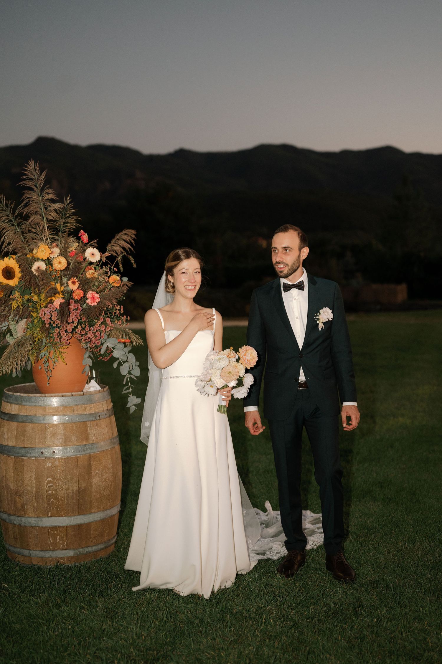 A couple in wedding attire poses near rustic wine barrels decorated with wildflowers at dusk in a garden setting.