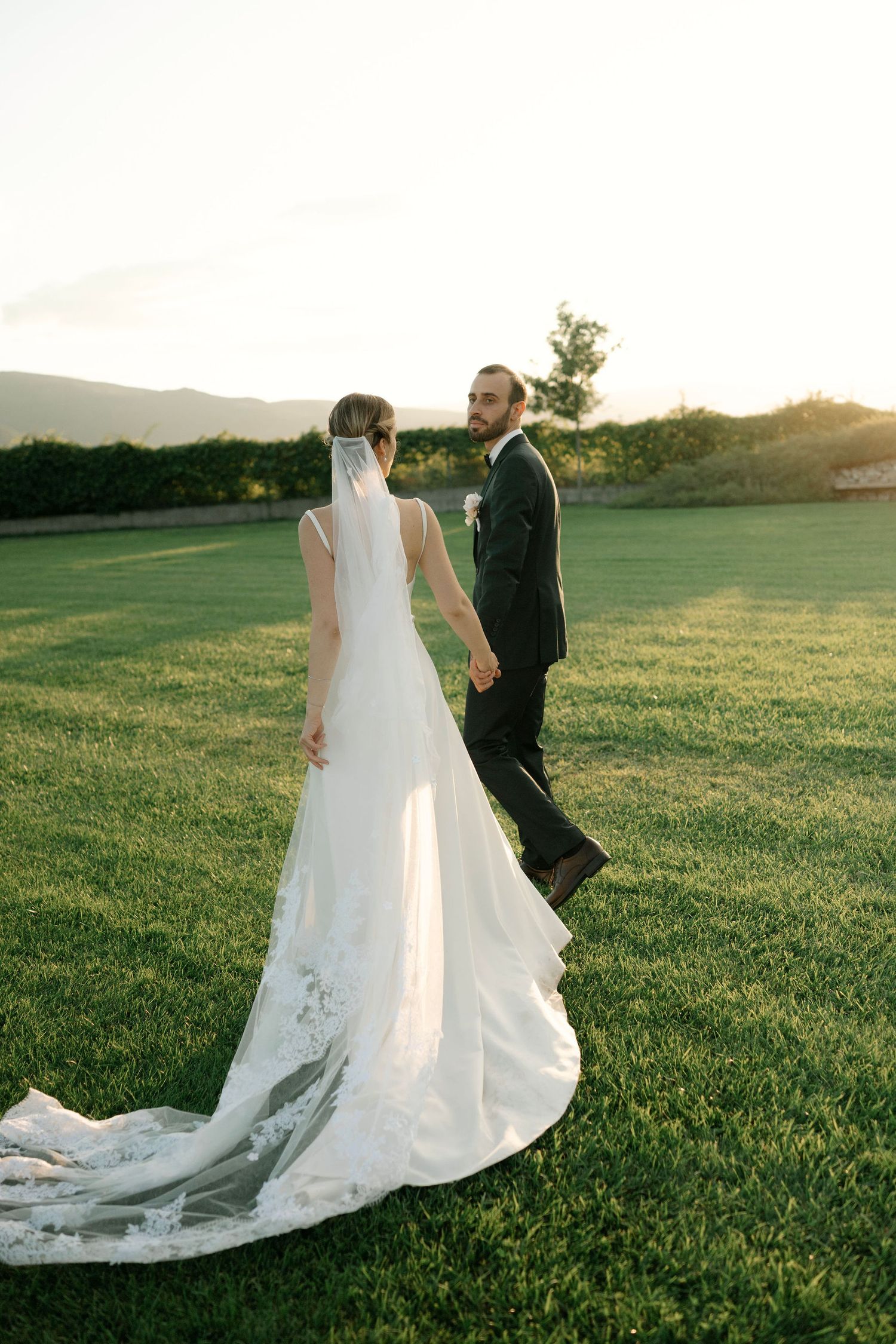 A bride and groom walk together on a lush green hillside at sunset as her long white train trails behind them.