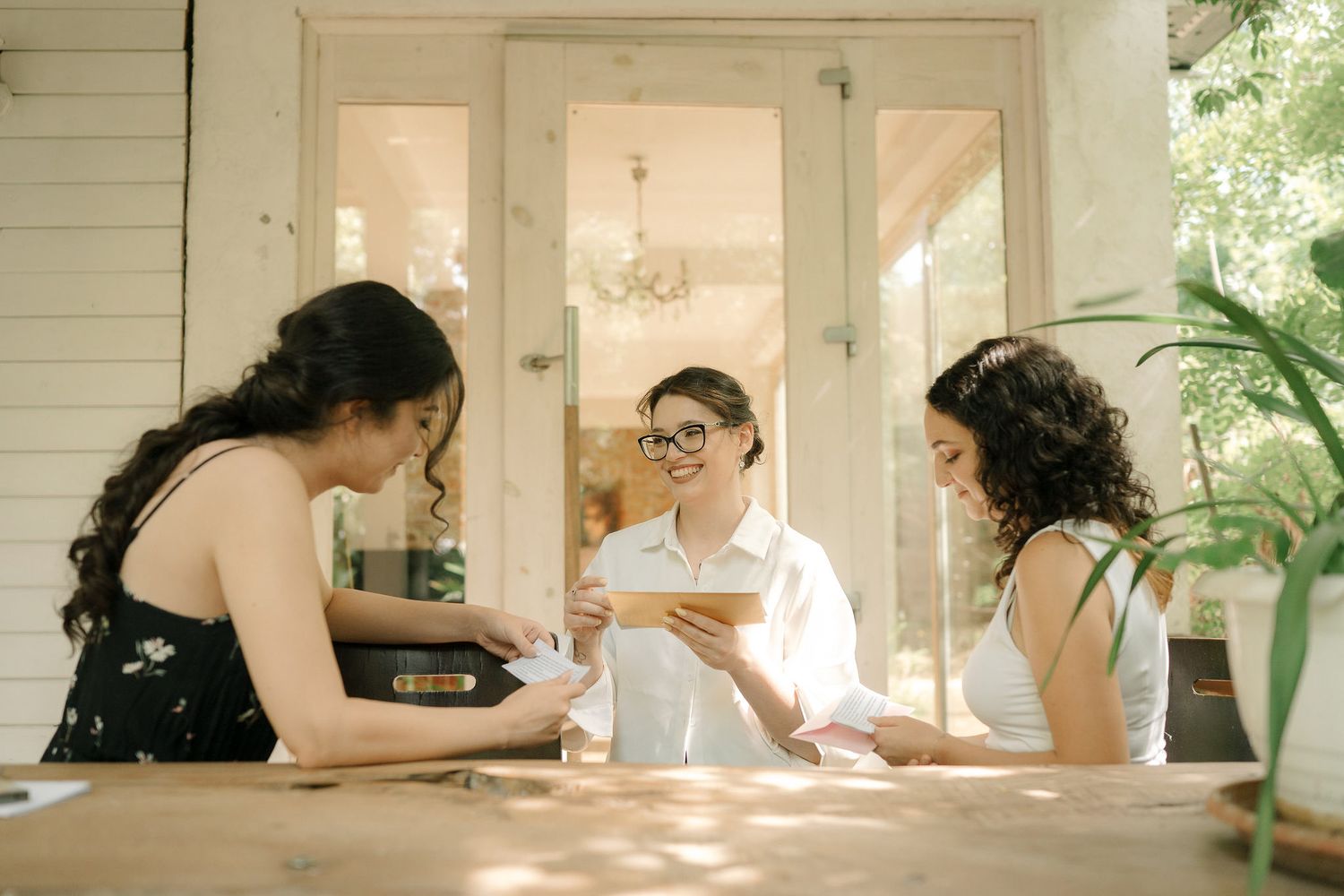 Three people sit at a wooden table having coffee and conversation in a bright, sunlit room with indoor plants.